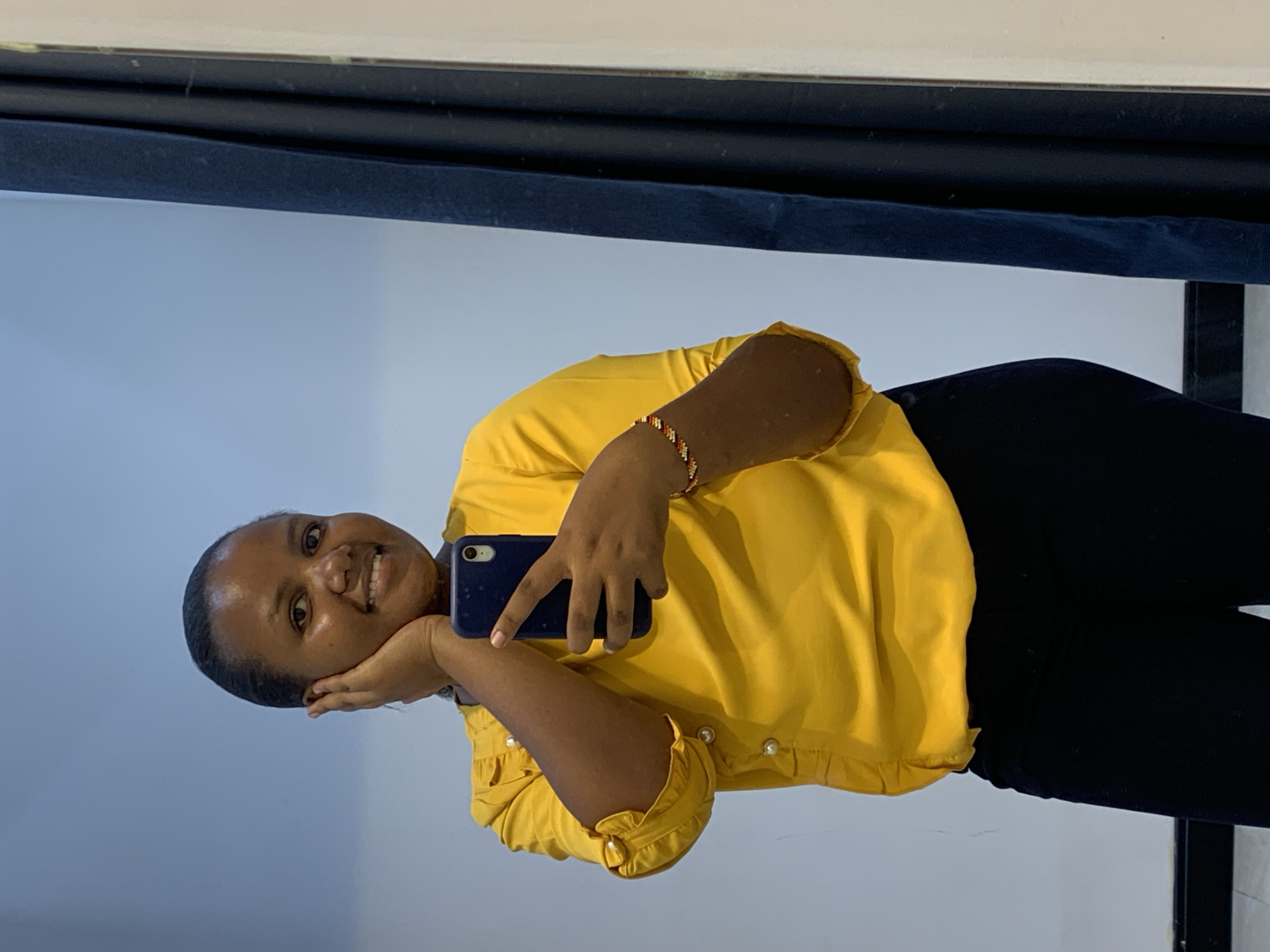 A woman with dark skin, wearing a yellow blouse with ruffled sleeves and black pants, is holding a blue smartphone and taking a selfie. She is smiling. She is wearing a beaded bracelet. She appears to be indoors with a dark-colored curtain or drape above her and a white or light-colored wall behind her. There is no visual information available to determine if the location is in Dar es-Salaam, Tanzania.