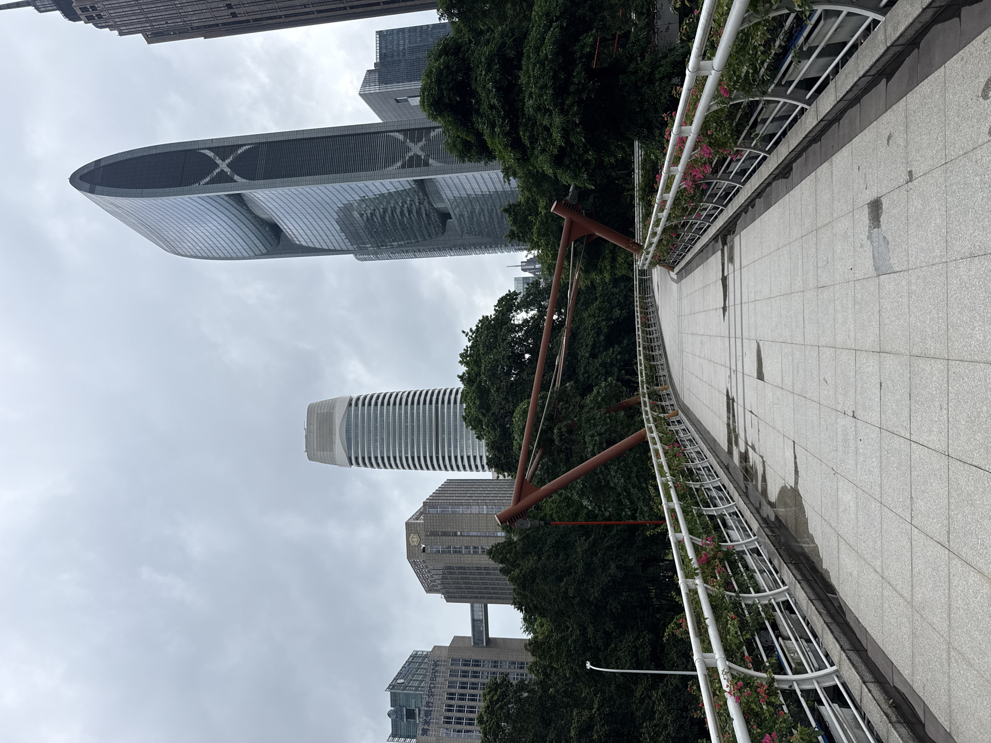 The image shows a pedestrian bridge in Tianhe District, China, with modern skyscrapers in the background. The bridge has a white railing adorned with pink flowers in planters. The walkway is made of light-colored tiles, with some wet patches indicating recent rain. The sky is overcast with gray clouds. Several tall buildings are visible, including one with a distinctive curved shape and an "X" design, and another cylindrical building with horizontal bands. Green trees surround the base of the buildings and are visible behind the bridge railing. The scene appears to be an urban landscape with a focus on architecture and infrastructure. There are no people visible in the image.