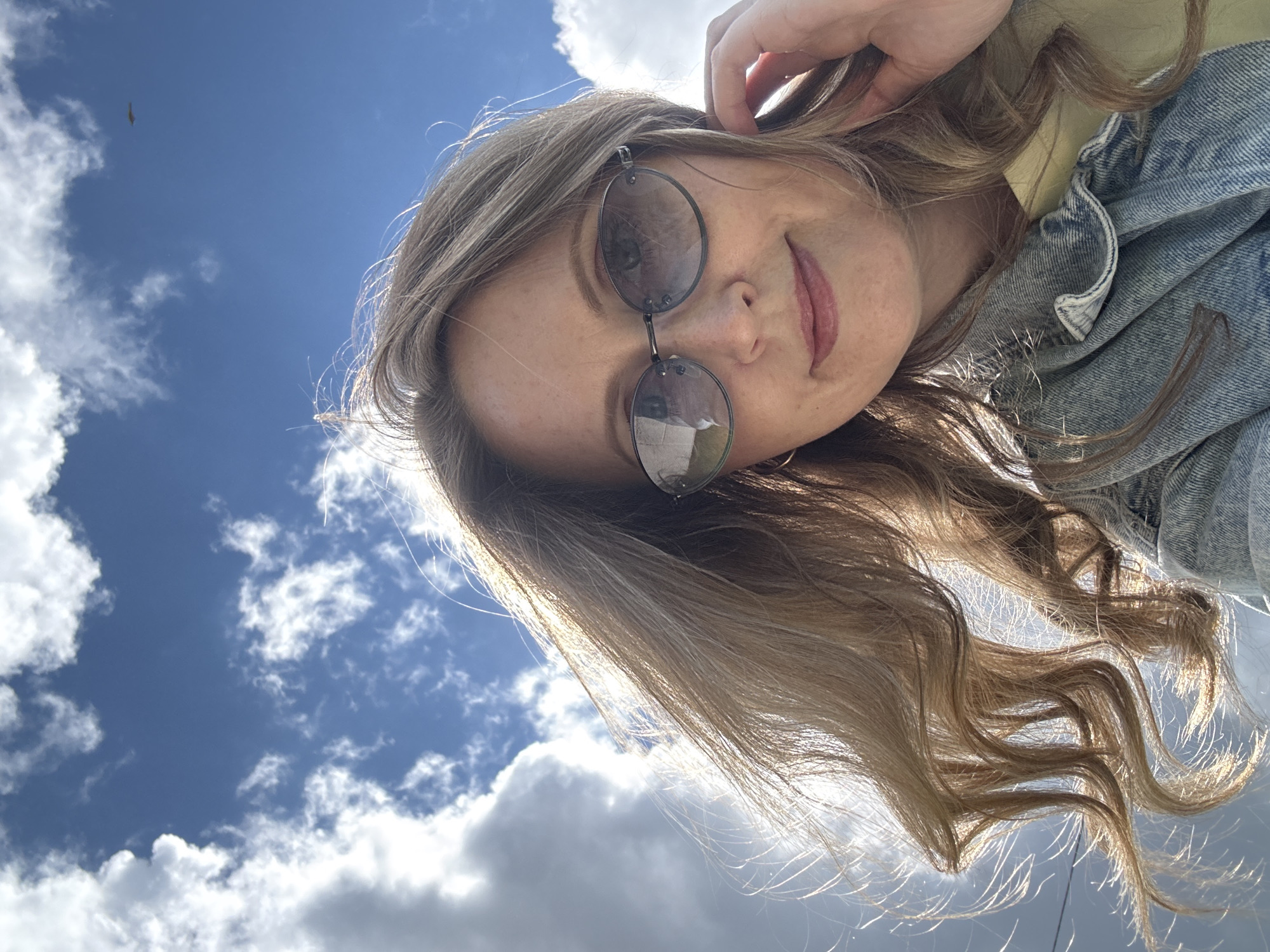 A young woman is taking a selfie outdoors against a backdrop of a bright blue sky with scattered white clouds. She is tilted to the right of the frame and is looking down at the camera through round-framed sunglasses. Her hand is raised towards her head. She appears to be wearing a denim jacket over a light-colored garment. The image has a bright, sunny feel. The location is Chaikovsky, Russia.