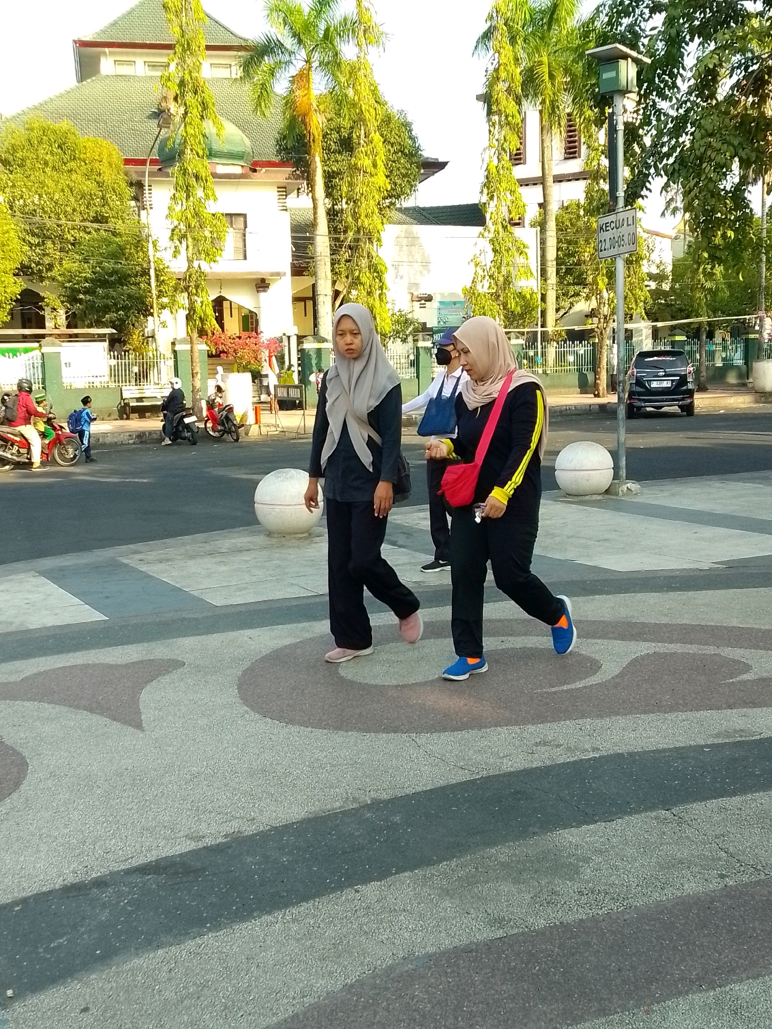 This image shows two young women wearing hijabs walking across a pedestrian crossing in Jember, Indonesia. The woman on the left wears a grey hijab and a dark blue long-sleeved shirt and black pants. The woman on the right wears a light brown hijab, a black long-sleeved shirt with yellow and white stripes on the sleeves, and black pants. She carries a red bag across her body. In the background, a black SUV is parked on the right side of the street, and a sign with Indonesian text is mounted on a pole. Behind the SUV and pole, a building with a green roof and white walls is visible. There are palm trees and other foliage throughout the scene. The pedestrian crossing has a patterned surface of grey and lighter colored shapes. Other people and motorcycles are visible in the background on the sidewalk and street. The scene appears to be a street in a public area.
