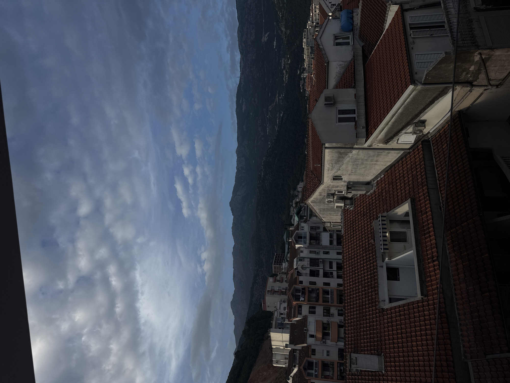 The image is a landscape of Budva, Montenegro. The foreground contains buildings with red tile roofs, apartment buildings, and air conditioning units. Beyond the buildings is a mountainous terrain extending from the bottom to the top center of the frame. To the left is a sky filled with clouds.