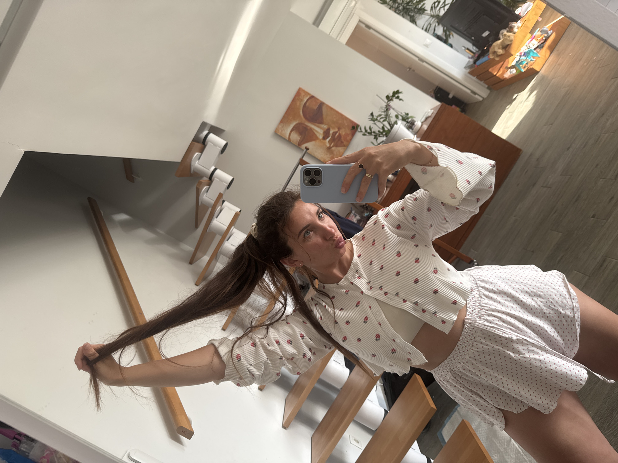 A young woman is depicted taking a mirror selfie within a brightly lit, modern home, presumably in Valencia, Spain. She holds a light blue-cased smartphone in her right hand, positioned to capture her reflection. Her long, dark hair is styled in two pigtails, and she playfully holds a section of her hair with her left hand while making a "pouty" or "kissing" face towards the camera.
She is dressed in a casual yet stylish outfit, featuring a white, ribbed button-down shirt or cardigan adorned with a repeating red/pink strawberry pattern, worn open over a white crop top. Her lower body is clad in light-colored shorts with a subtle dotted pattern and an elasticated waist. A gold ring with a dark stone is visible on a finger of her right hand.
The background, reflected in the mirror, reveals a clean, contemporary interior. White walls dominate the space, complemented by light wooden elements such as stair treads or shelves. On the wall to her left, several modern dispensers hold white cylindrical objects, possibly toilet paper rolls. Further into the room, a warm-toned abstract painting hangs on a wall, and a green plant adds a touch of nature. Beyond that, a television or monitor is visible, along with a wooden shelf unit displaying various items. The floor features light wood-look tiles. The ample natural light indicates it is daytime. The overall atmosphere is lighthearted and playful. No discernible text is visible in the image.