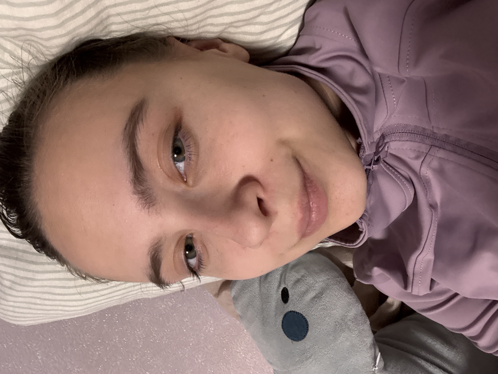 A woman with fair skin and dark hair is lying on a bed with striped sheets. She is wearing a purple zip-up jacket. A grey stuffed animal is next to her. The woman's eyes are open and she has a slight smile. The location is unspecified, and there is no indication of it being in Прикамский, Russia.