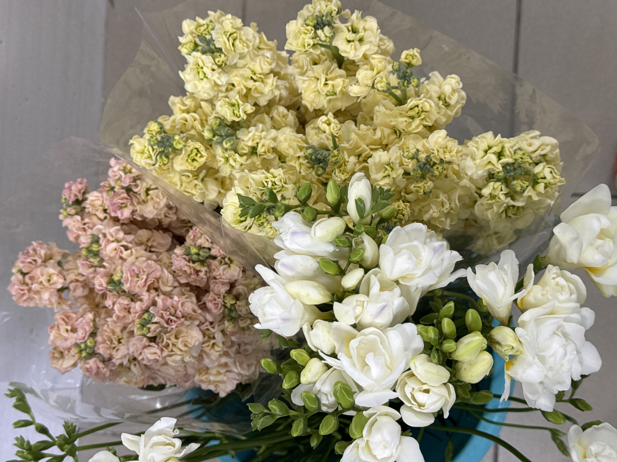 The image shows three bouquets of flowers, likely for sale, in what appears to be a retail setting. Two of the bouquets are densely packed with small, ruffled flowers in soft peach and pale yellow hues. The third bouquet, to the left and foreground, consists of white freesia flowers, characterized by their delicate, trumpet-shaped blooms and slender green stems. The flowers are wrapped in clear cellophane, some tied with string or rubber bands. The background is a neutral, textured wall, and a light-colored floor is visible at the bottom. The overall impression is one of a floral arrangement display. The image does not contain any specific geographical markers to identify its location within Izhevsk, Russia.