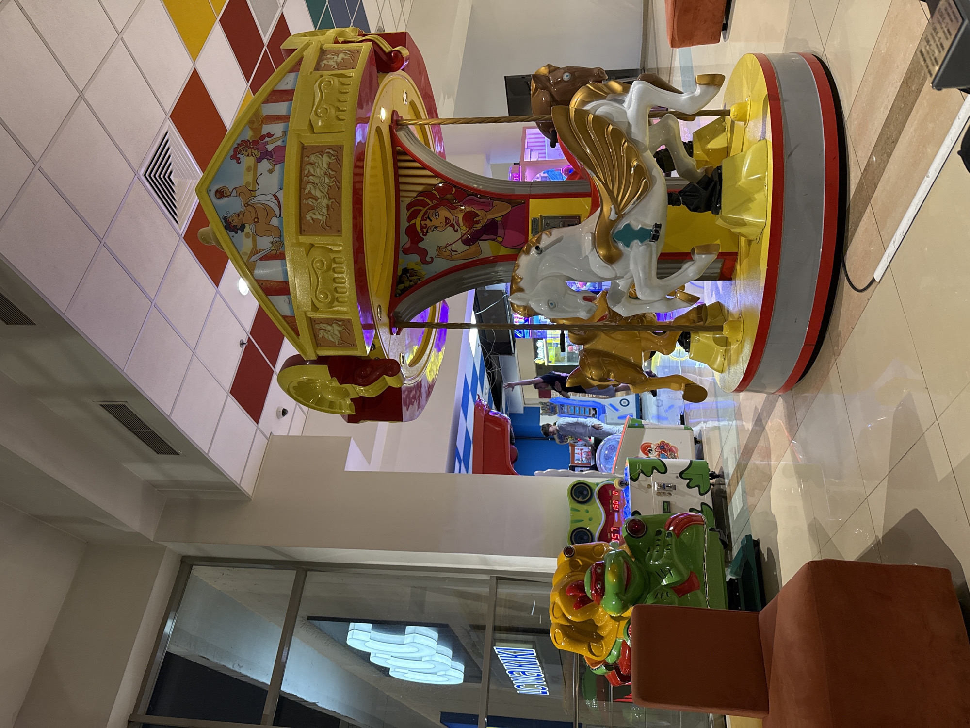 The image depicts an indoor amusement area featuring a carousel with horse figures and cartoon decorations. The scene includes arcade-style games and seating, with a visible window reflecting interior lighting. The location is identified as Icmeler Turkiye.