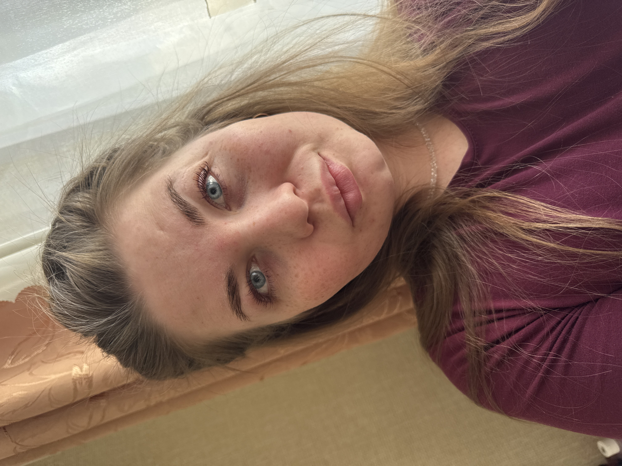 The image is a selfie of a young woman with fair skin, freckles, and light brown hair, posing indoors. She has blue eyes and is wearing a dark red top. A delicate necklace is visible. The background appears to be a wall or surface in shades of beige and cream, with a light-colored curtain or drapery visible near the top of the frame. The image is taken at an angle, with the woman's head tilted. The location within the city of Chaikovsky, Russia is unknown.