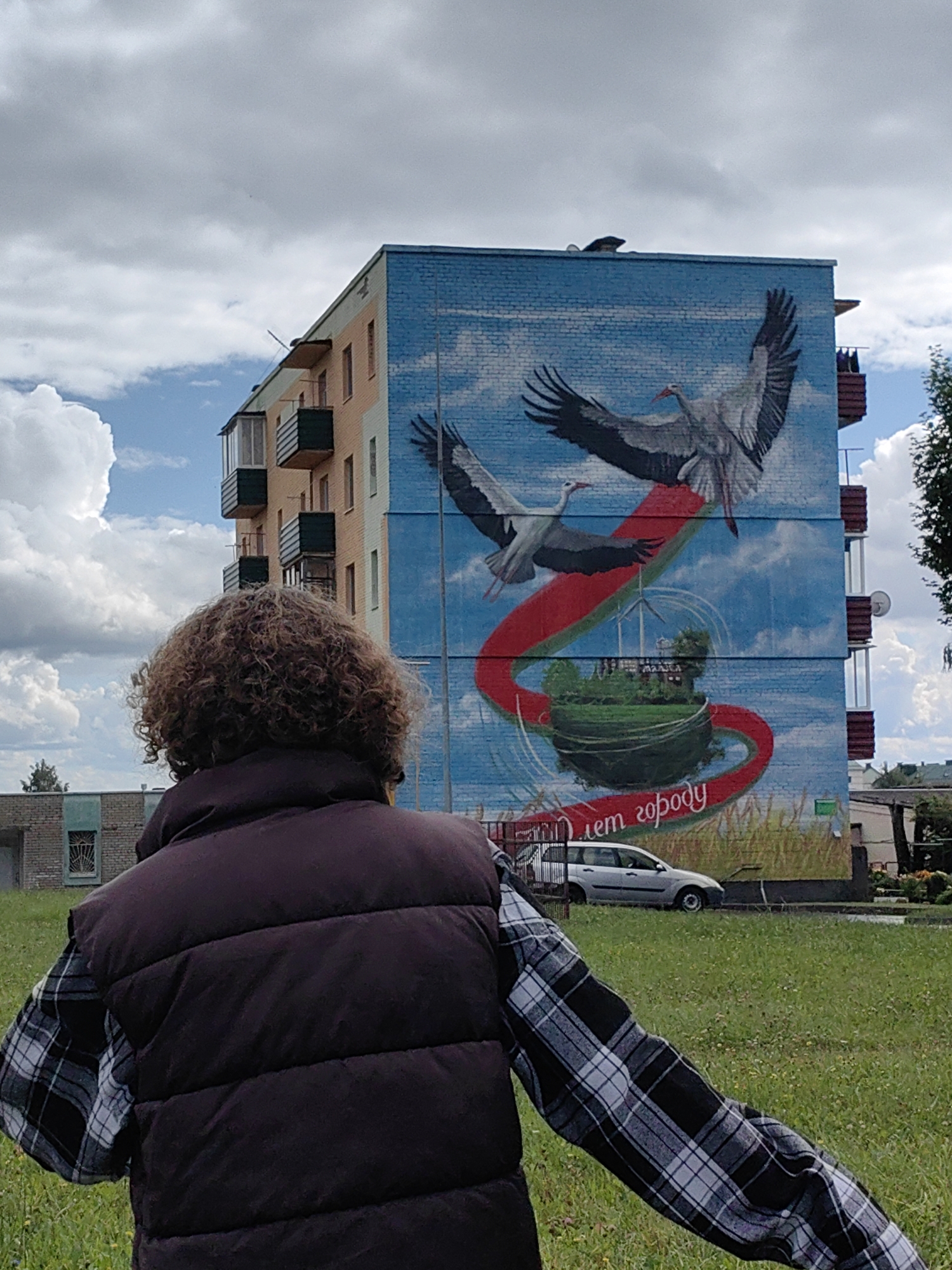 In Myadzyel, Belarus, a person with curly brown hair, viewed from behind, stands in a grassy area facing a large mural on the side of a brick apartment building. The mural depicts two storks in flight against a blue sky with clouds. Below the storks, a red ribbon curves around a green circular landscape with a wind turbine and buildings, and text in Russian that appears to say "С днем города" (Happy City Day). A silver station wagon is parked in front of the mural, and the sky is cloudy with patches of blue. The person is wearing a dark puffer vest over a black and white plaid shirt. The environment is residential, with other buildings and trees visible in the background.