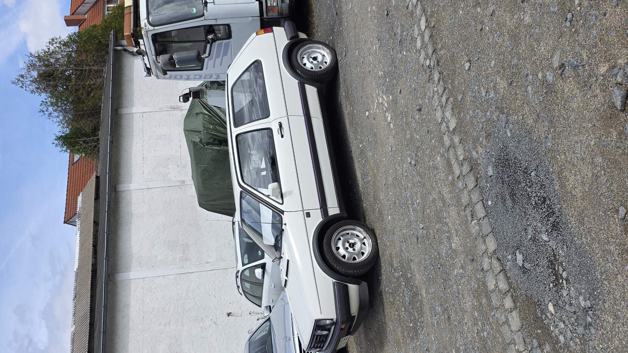 A white Volkswagen Golf MK2 hatchback is parked on a gravel lot. The car is a two-door model and appears to be in good condition, with clean bodywork and intact wheels. The car is parked next to other vehicles, including a silver Audi A3 and a large white truck. In the background, there is a white building with a corrugated roof, and beyond that, houses with red-tiled roofs and trees. The sky is partly cloudy. The overall scene suggests a parking lot or storage area, possibly in an industrial or rural setting.