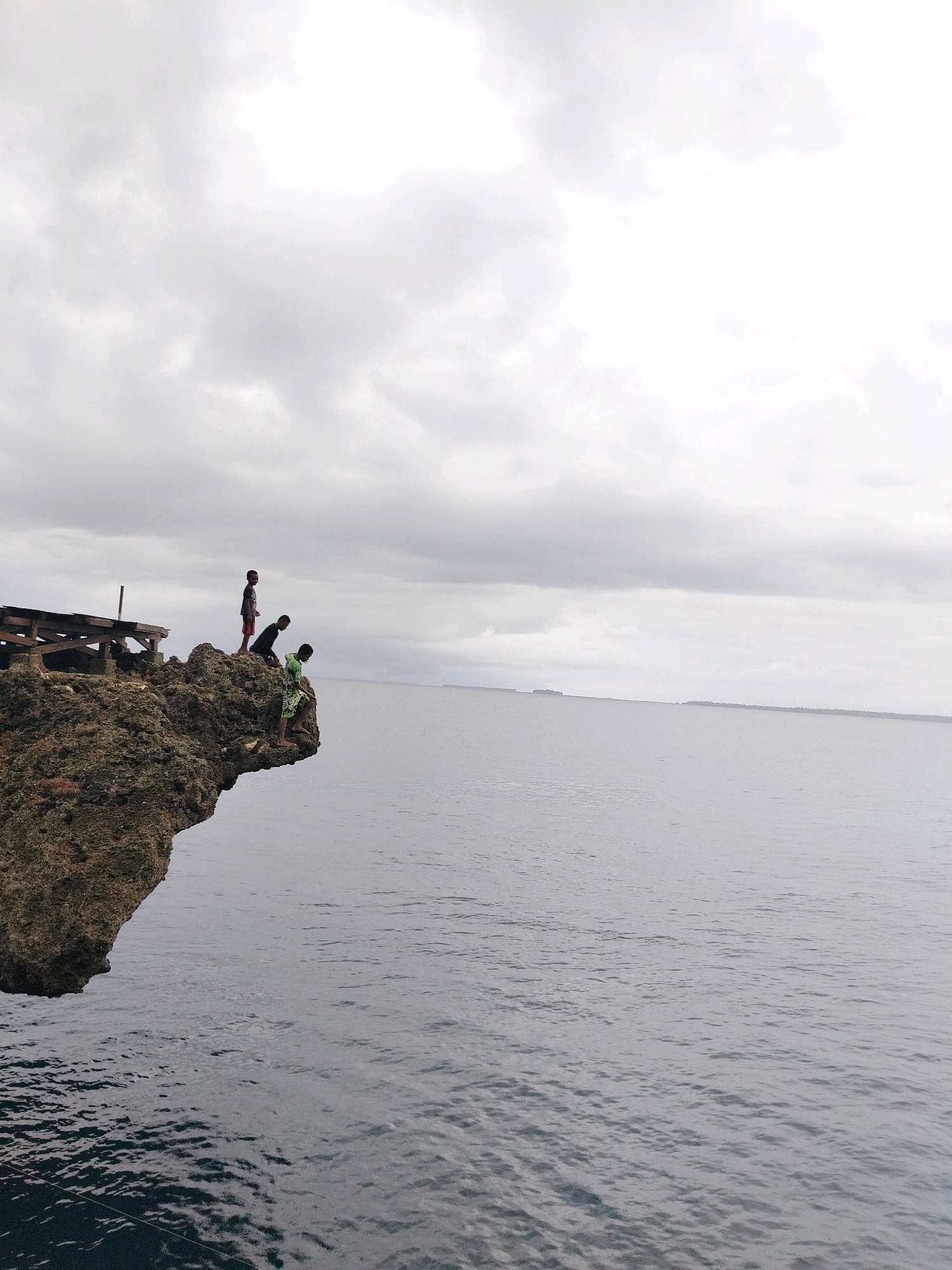 This is a medium shot of three boys standing on a rocky cliff overlooking the ocean. The cliff is rough and brown, with some vegetation growing on it. There is a wooden structure on the cliff to the left of the boys. The ocean is calm and grey, with a distant landmass visible on the horizon. The sky is overcast with grey clouds. The boy on the left is wearing a dark t-shirt and red shorts. The middle boy is wearing a black shirt and dark shorts. The boy on the right is wearing a green and white patterned shirt and green shorts. He is leaning over the edge of the cliff. The location is the cliffs of Animi, Indonesia.