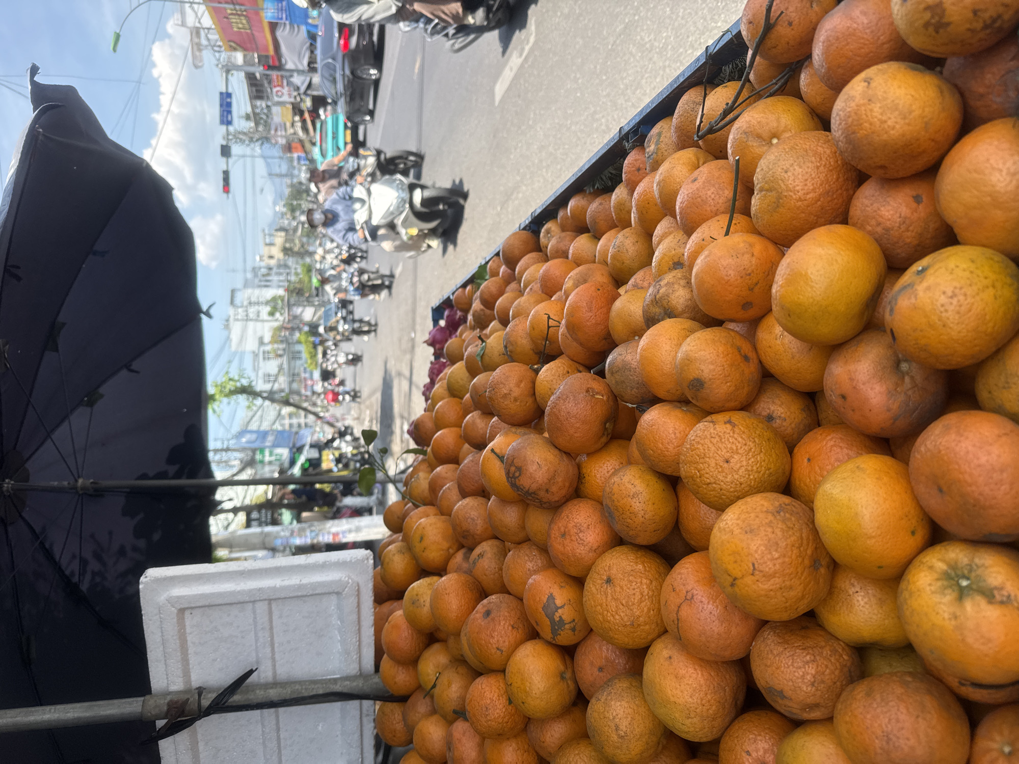 The photograph shows a large pile of oranges filling the right side of the frame. To the left, a black umbrella and a white container are visible. In the background, a busy street scene unfolds with motorbikes, cars, pedestrians, and buildings. The location appears to be a street in Nha Trang Ward, Vietnam, suggested by the architecture and traffic patterns. The image captures a street-side fruit vendor.