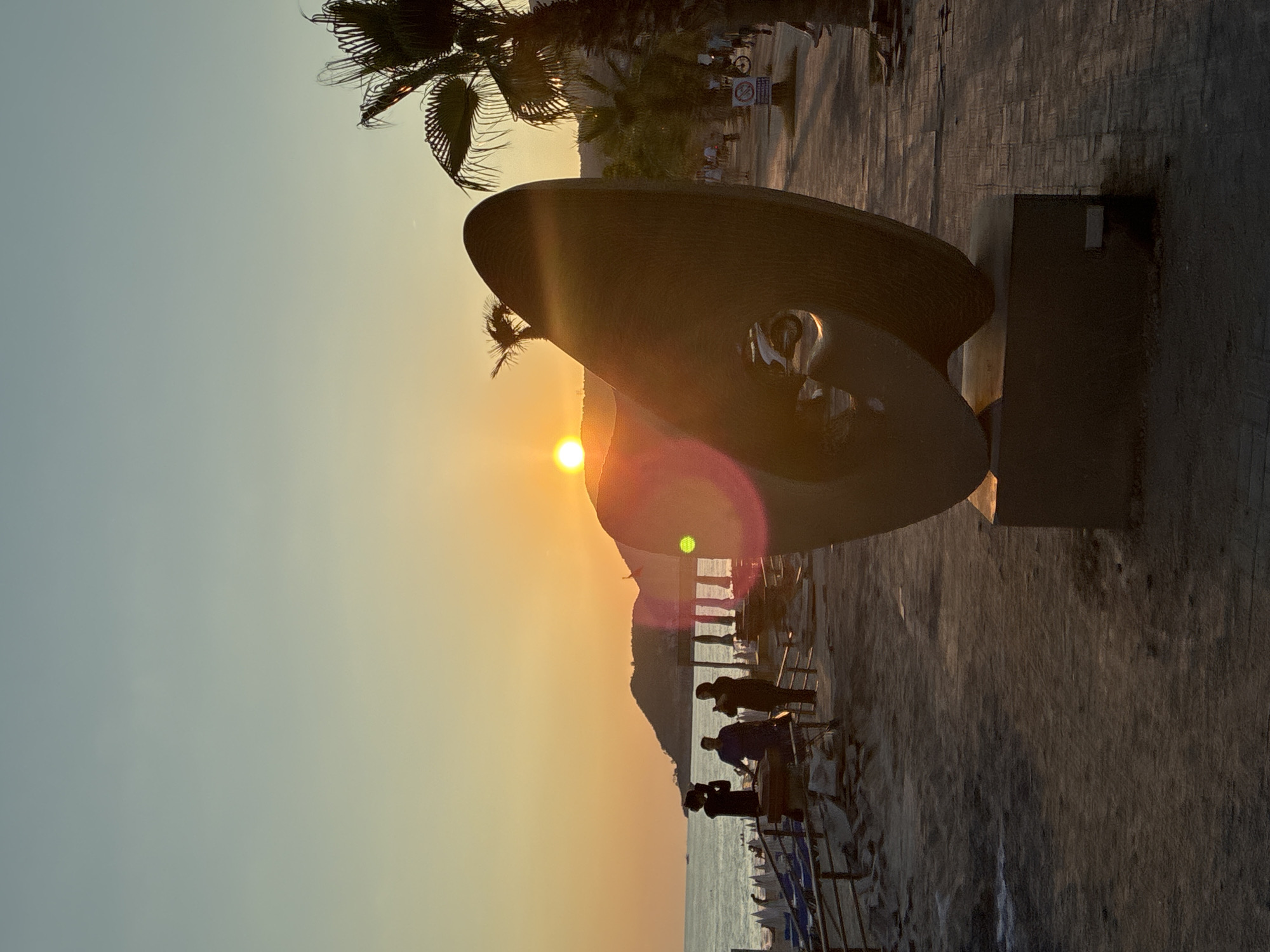 The image depicts a sunset scene on a beach promenade in Alanya, Turkey. A large, abstract sculpture stands in the foreground, casting a shadow on the paved walkway. To the left, the ocean is visible, with a coastline and hills in the distance. Several people are silhouetted against the setting sun, some appearing to be taking photographs. Beach chairs and umbrellas are arranged on the sand. Palm trees line the promenade, and a few other individuals are walking or cycling further down the path. The sky is a gradient of orange and pale blue, characteristic of a sunset. A lens flare is present in the lower right portion of the image.