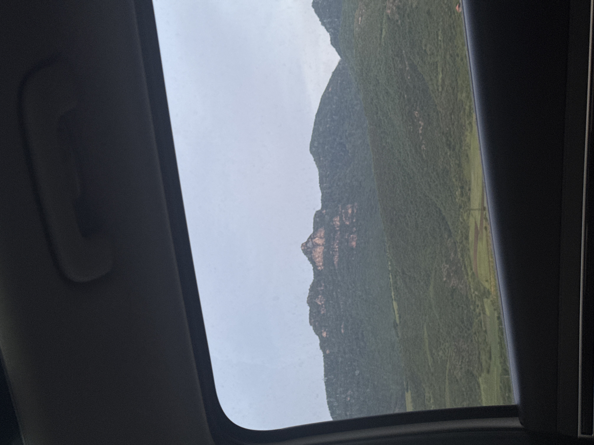 The image is taken from inside a car, looking through an open sunroof at a mountain landscape. The sky is overcast and gray. The mountains are covered in dense green vegetation. The location is Unknown, Armenia. The primary subject is the landscape seen through the car's sunroof.
