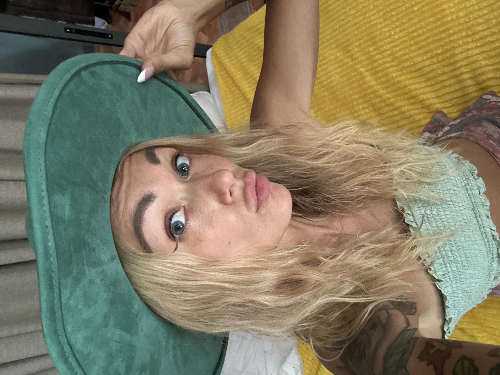 A blonde woman wearing a green hat and a light blue top is lying on a yellow textured surface. She is making a duck face with her lips. She has tattoos on her arm and shoulder. The background appears to be indoors with curtains and some furniture. There is no indication of the city of Mũi Né, Vietnam in this image.
