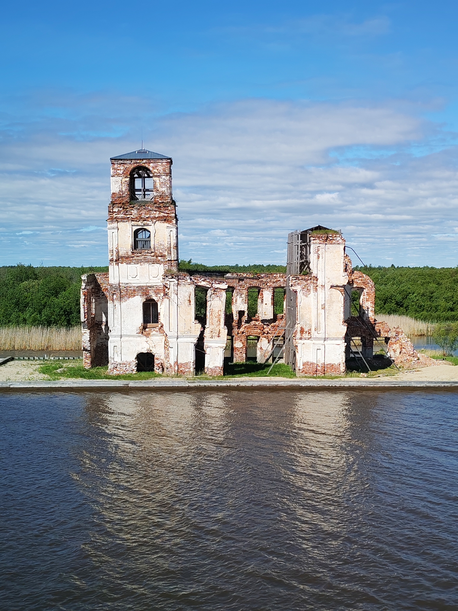 The image shows the ruined Church of the Nativity in Krokhino, Russia. The church is standing in water, likely part of the Sheksna River or related reservoir. The building appears to be made of brick and white plaster. The building has a prominent tower with a small roof structure on top. The walls are largely missing, exposing the brick interior. Some scaffolding is visible on the side of the building. A small strip of land surrounds the church, separating it from the water. The sky is blue with scattered white clouds. Lush green trees are visible in the background. The water is dark and reflects the building.