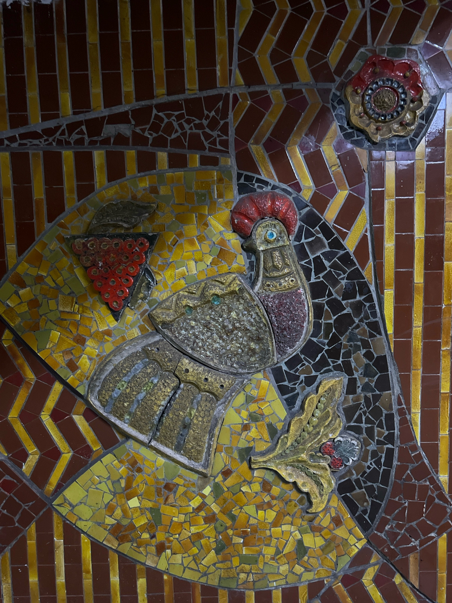 The image displays a close-up of a decorative mosaic tile artwork depicting a stylized rooster or bird, a cluster of red berries resembling grapes, and a floral element. The mosaic is set against a background of vertical reddish-brown and yellow tiles arranged in alternating patterns, with some areas filled with small, irregularly shaped mosaic pieces in shades of yellow, green, and brown. The central subject is a golden-toned metallic bird with a red crest and a blue eye, detailed with what appears to be textured metal for its body and wings. A cluster of red, circular mosaic pieces forming grapes is positioned to the upper left of the bird. A golden-brown leaf-like motif with small red berries and a central blue and gray element is located to the lower right. This mosaic artwork is part of the decorative elements found on the walls of the Kyiv Metro system. Specifically, this mosaic is located in the Zoloti Vorota (Golden Gate) station.