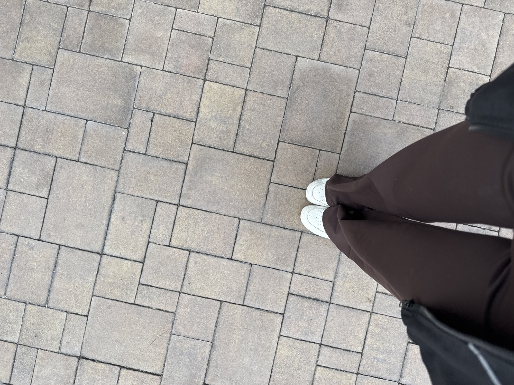 An overhead shot captures a person standing on an outdoor paved surface in Rostov-on-Don, Russia. The lower half of the person's body is visible, showcasing their feet in clean white sneakers with perforated detailing on the sides. They are wearing dark brown, wide-leg pants that drape over the shoes. A portion of a dark-colored bag or jacket is visible on the right side of the frame, suggesting it's slung over the person's arm or shoulder.
The ground is covered with an intricate pattern of rectangular and square concrete pavers. These pavers are primarily in muted tones of grey and light beige, with dark, well-defined grout lines separating each block. The pattern appears somewhat irregular, with different sized blocks intermingling. The surface texture of the pavers suggests a slightly rough, stone-like finish.
The lighting indicates it is daytime, with soft shadows cast by the person's legs, suggesting either an overcast sky or indirect sunlight. The pavement appears dry. No other subjects, distinct objects, or visible text are present in the image. The scene conveys a casual moment in an urban or suburban outdoor environment.