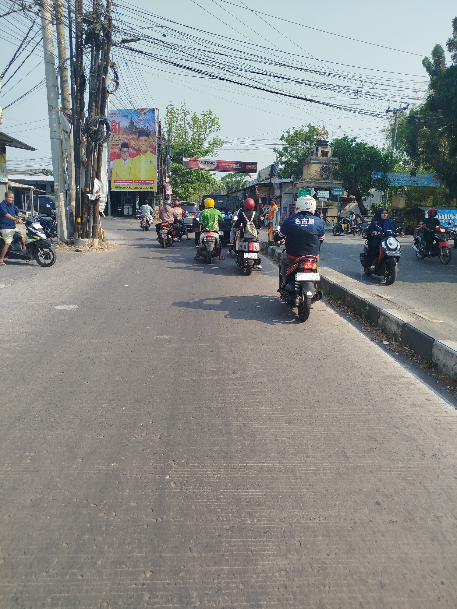 The image depicts a street scene in Semarang, Indonesia, likely a busy urban area characterized by significant overhead electrical wiring and a mix of vehicles. In the foreground, a gray asphalt road with visible tire tracks and some imperfections occupies the majority of the frame. Several motorcycles are moving in the same direction on the road.
On the left side of the road, a man in a blue shirt and shorts is standing next to a black scooter. Further up the road, a billboard with images of two men in yellow shirts and the text "INDONESIA MAJU" is prominently displayed, suggesting a political campaign or event. Adjacent to the billboard, a cluster of utility poles with a dense network of cables is a prominent feature of the urban environment.
Several individuals are riding motorcycles:
- A person in a green shirt and yellow helmet on a scooter is visible in the middle ground.
- Behind them, a person wearing a red helmet and a backpack on a scooter.
- Further back, a person in a blue jacket with Japanese characters (名古屋 - Nagoya) on the back is riding a motorcycle.
- To the right of this individual, a woman in a blue headscarf is riding a scooter.
- Further down the road, another motorcycle rider in a black helmet is visible.
In the background, there are buildings and trees, with a notable arched structure with an ornamental top. The sky is clear and bright, indicating daytime. The presence of numerous motorcycles and overhead wires suggests a typical streetscape in a bustling Indonesian city. The general impression is one of active daily life and transportation.