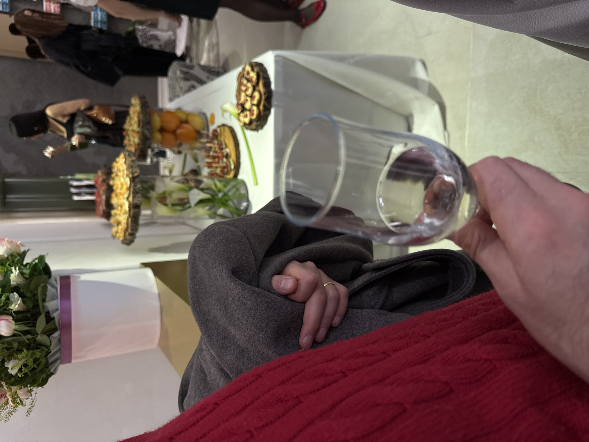 The image captures an indoor gathering, potentially a reception or celebration, in Yerevan, Armenia. In the immediate foreground, a person's right hand holds a clear glass containing water, positioned above a textured red fabric, possibly a blanket or throw. The person is wearing a gray coat or jacket, and a simple gold band is visible on their ring finger.
The background reveals a bustling scene centered around a long table laden with an assortment of food. Various dessert-like items or small appetizers, presented on rustic wooden platters, are visible, including round tarts topped with berries and other colorful toppings. A bowl filled with citrus fruits, likely oranges or mandarins, is also part of the spread. Decorative white calla lilies in a vase adorn the food table, and further to the left, a vibrant bouquet of pink and white flowers, possibly roses or tulips, sits in a white and pink cylindrical vase.
Several blurred figures of people are seen in the midground and background, milling around the food display. One woman with dark hair, dressed in a dark outfit, is partially visible near the table. Another person's lower legs and feet, wearing distinctive red shoes, can be seen further back. The overall lighting suggests an indoor setting with artificial illumination. No visible text is present in the image. The scene conveys a social event with a focus on refreshments and conversation.