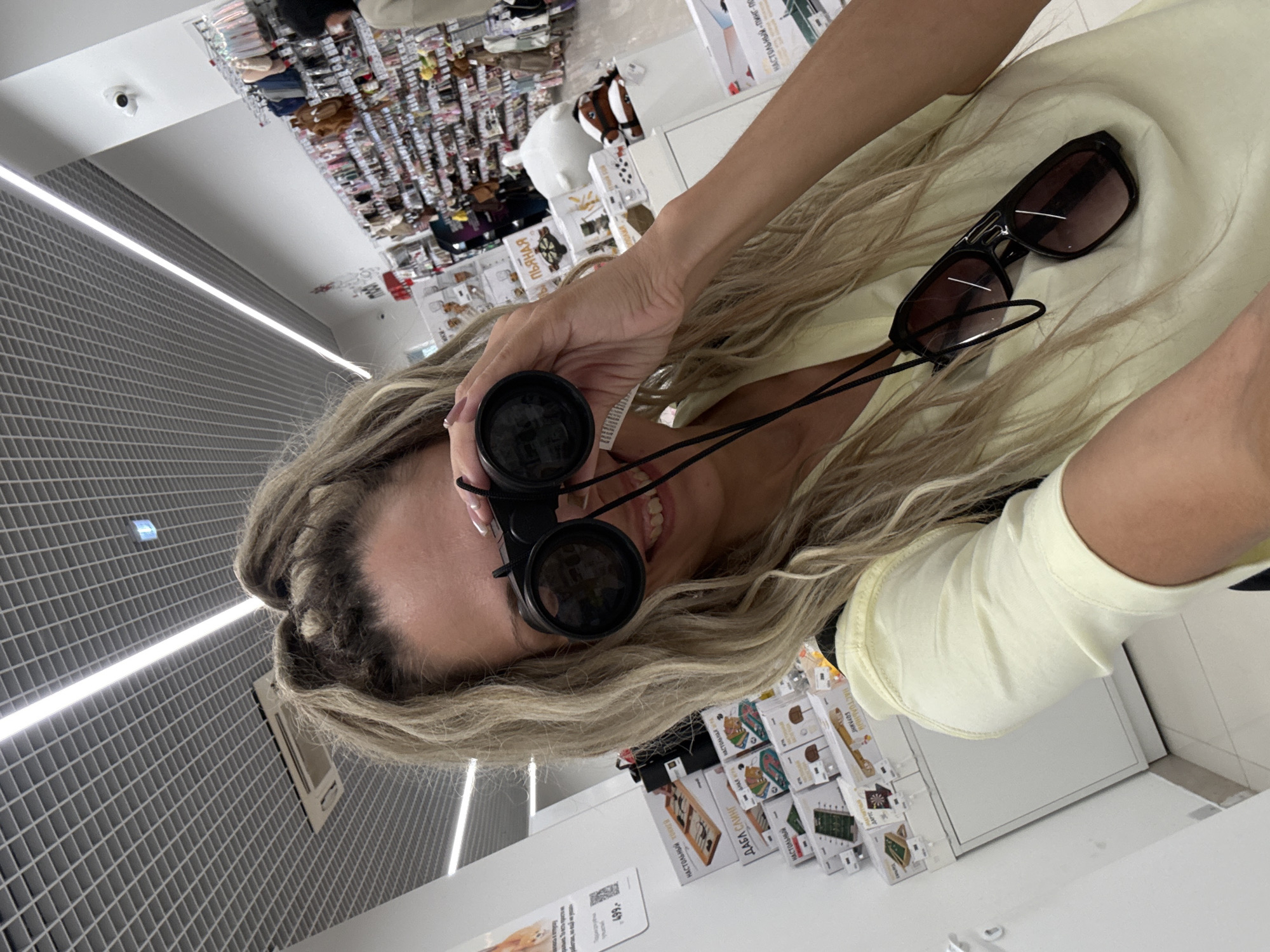 A young woman with blonde hair styled in braids is taking a selfie in a retail store. She is holding a pair of black binoculars up to her eyes, looking through them. She is wearing a light yellow t-shirt, and a pair of black sunglasses are hanging from a cord around her neck. The background of the image shows shelves stocked with various products, suggesting a retail environment. The ceiling is a grid of white acoustic tiles with fluorescent lighting. The location is identified as a store in Sosnovtsy, Russia.