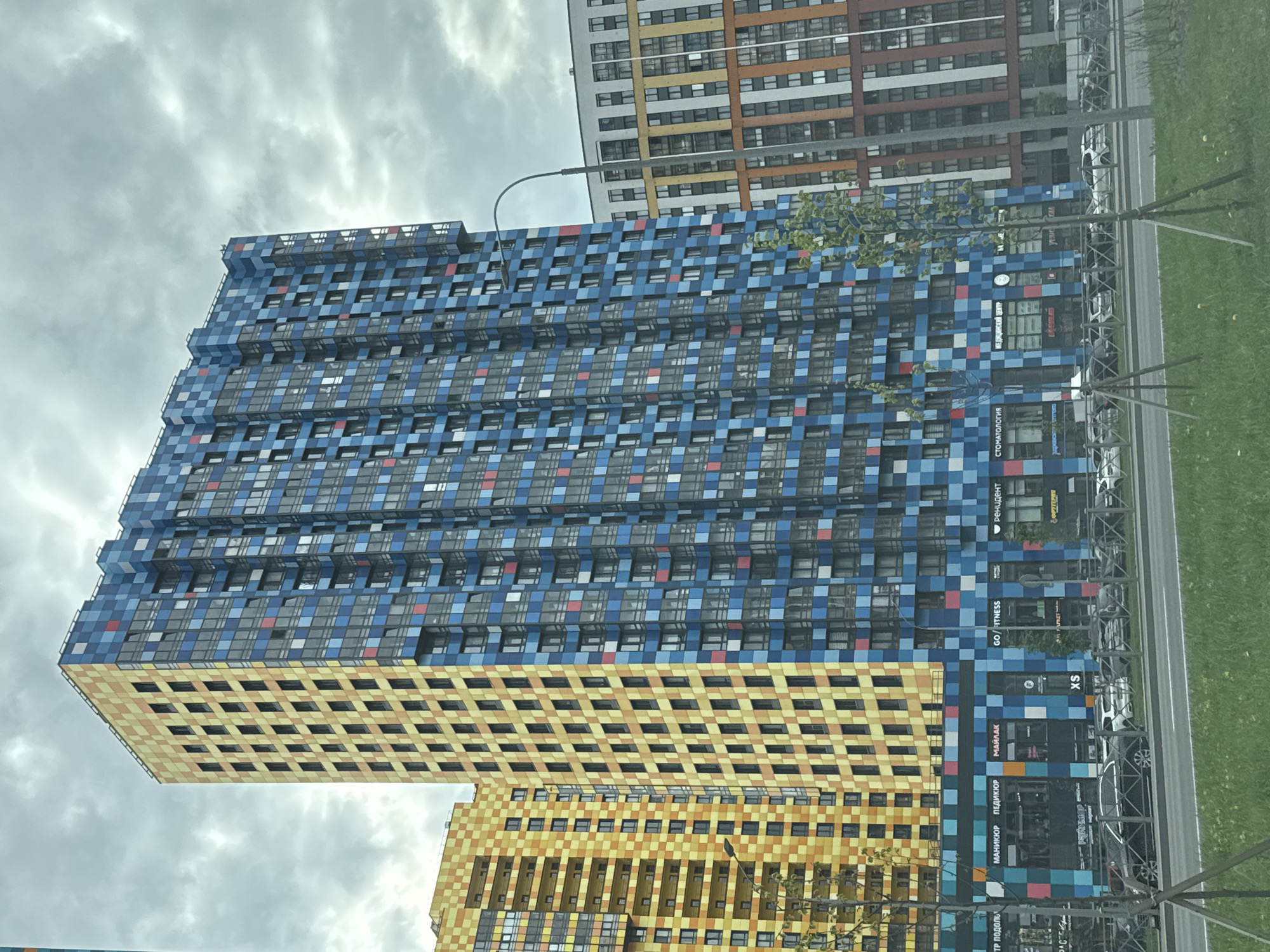 The image shows a tall, modern apartment building in Saint Petersburg, Russia. The building's facade is characterized by a mosaic-like pattern of blue, yellow, and white squares, giving it a distinctive appearance. Windows are embedded within the mosaic design. At the base of the building, there are several businesses with signs in Russian and English, including a fitness center (GO/FITNESS), a nail salon (МАНИКЮР ПЕДИКЮР МАЙЛАК), a medical center (МЕДИЦИНСКИЙ ЦЕНТР), a dental clinic (СТОМАТОЛОГИЯ) and a supermarket (ФРУТЕРИЯ).
A street runs alongside the building, with parked cars and a narrow strip of grass. In the background, another tall apartment building can be seen, with a vertically striped facade in various shades of orange, yellow, white, and gray. The sky is overcast with gray clouds. The photograph is taken from a low angle, looking up at the buildings.