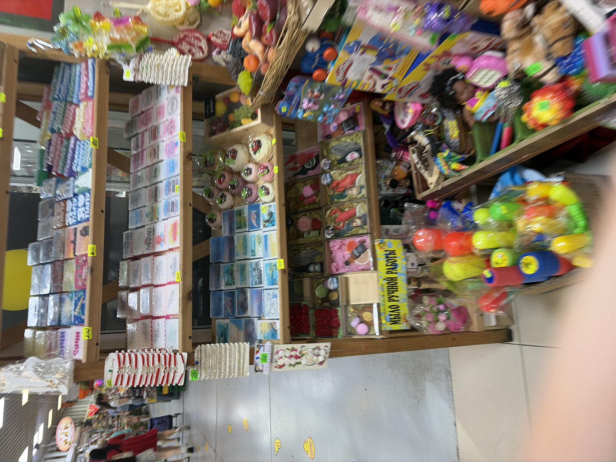 This is a photograph of a market stall displaying various goods, likely in Sochi, Russia, given the visible text. The stall is packed with items, including small packaged soaps, jewelry, toys, and what appear to be decorative items like artificial flowers. The overall scene is colorful and somewhat cluttered, suggesting a bustling retail environment.
The photograph captures a wide array of merchandise, organized on wooden shelves. Prominently featured are rows of small, individually wrapped items, many of which are labeled in Russian. Some text visible includes "Сочи" (Sochi) and "МЫЛО РУЧНОЙ РАБОТЫ" (handmade soap), confirming the location and type of some products. Other labels like "MAMA," "Дяди" (Uncle), and "БРАТА" (Brother) suggest the products might be marketed as gifts. There are also price tags visible, indicating prices like "100," "150," and "200."
Among the items, there are also displays of bracelets, toys such as colorful water guns or squirt toys, and plush toys. The lighting appears to be artificial, typical of an indoor market or a covered outdoor space. The image is slightly blurry in parts, particularly the background, and the angle suggests the photo was taken by a visitor or vendor. No people are clearly identifiable as subjects, though glimpses of figures might be present in the background, blurred by motion or distance. The setting is a retail space, likely a flea market or a dedicated souvenir shop, given the variety and presentation of the goods. The time of day is not discernible, but the artificial lighting suggests it could be any time.