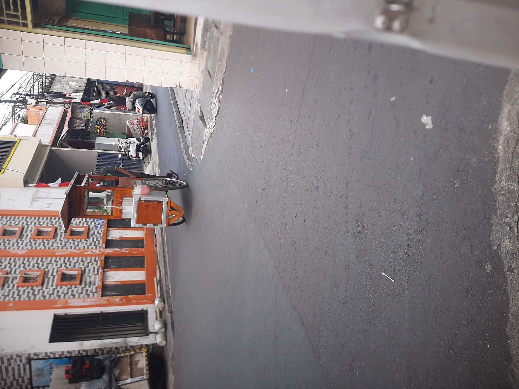 In Central Jakarta, Indonesia, an outdoor street scene is captured. A vendor, wearing dark clothing, is operating a food cart with a wooden top and metal base, pushed by a bicycle. The cart is positioned on the asphalt road. In the background, a white scooter is parked and two individuals are visible on a separate white scooter further down the street. Buildings line the street, one featuring an orange and gray patterned facade with rectangular window openings, and another with a yellow trim. Red and white flags are displayed on poles. The street is paved with asphalt and concrete sidewalks. Debris, including cylindrical objects and scattered paper, is present on the sidewalk. The lighting suggests it is daytime.