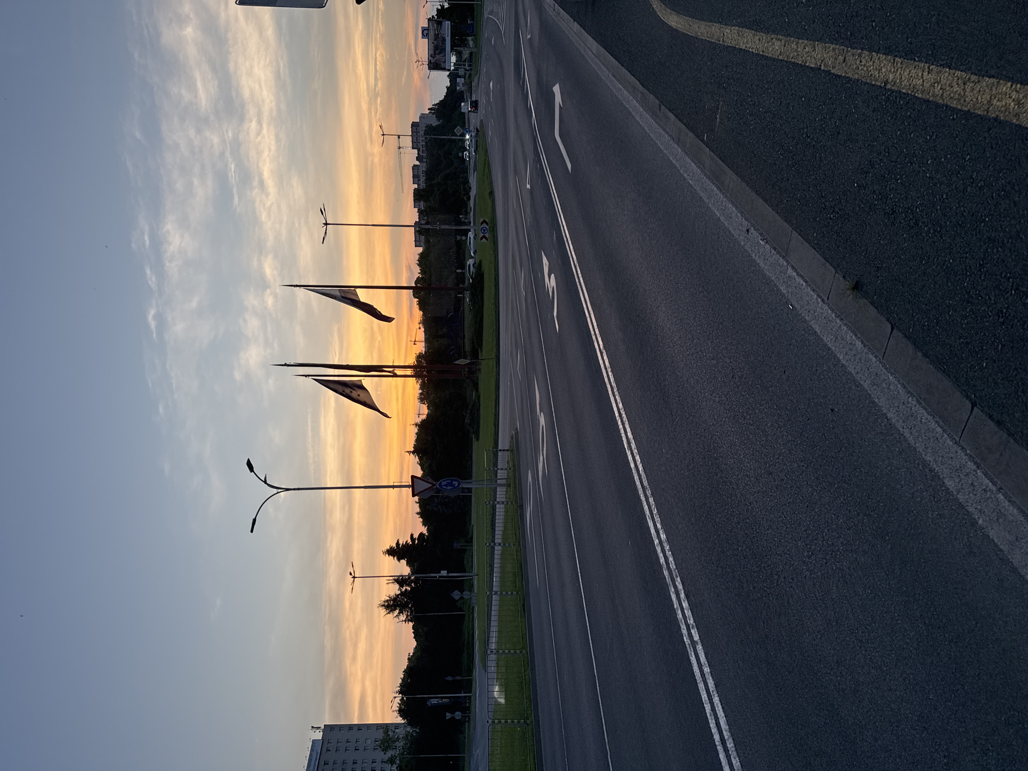 The image captures an urban landscape at either sunrise or sunset, evidenced by the sky's gradient transition from light blue at the top to vibrant orange and yellow hues near the horizon. Sparse white clouds are scattered across the sky.
In the foreground, a dark asphalt road surface displays double solid white lines in its center and two distinct white road markings: one indicating a straight path and another a right turn. A grey concrete curb delineates the road's edge on the far right, adjacent to a solid yellow line on a separate paved section.
The midground features a wide road or intersection. A metal barrier and a grassy area are visible on the left. Multiple streetlights are present, including a tall pole with two curved arms on the left and several multi-lamp fixtures further into the distance. Various traffic signs are identifiable: an inverted red triangle (yield), a blue circular sign with white arrows indicating a roundabout, and a blue rectangular directional sign. Several flags on tall poles are silhouetted against the sky, with one flag on the right appearing to have three stars. Dark, dense trees and bushes line the roadside and obscure parts of a central traffic island or roundabout. In the distance, several vehicles are observed on the road, some with illuminated headlights.
The background reveals a tall, multi-story building with numerous windows on the far left. Along the horizon, additional buildings and multiple construction cranes are silhouetted against the bright sky, suggesting urban expansion. No individuals are visible in the scene.