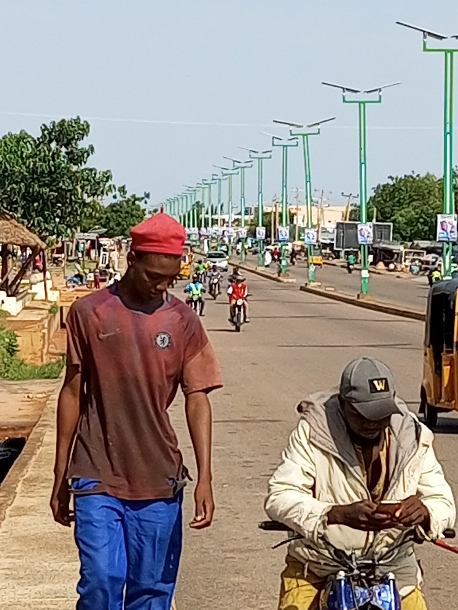 The image depicts a street scene in Tempure, Nigeria. In the foreground, two men are walking. The man on the left wears a red beret and a reddish-brown t-shirt with blue pants. The man on the right wears a grey baseball cap with a "W" logo and a light-colored jacket over a patterned shirt, with yellow pants. He appears to be looking at a mobile phone in his hands. Behind them, the street is lined with solar-powered streetlights and various businesses. Several people are on motorcycles and a three-wheeled vehicle is visible on the right. The environment is sunny with a clear sky.