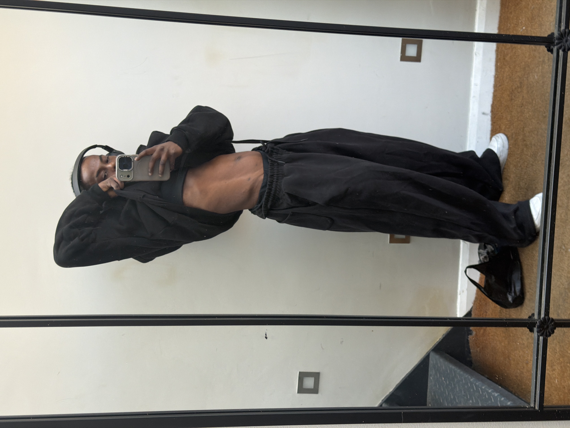 An individual with dark skin, wearing black over-ear headphones, takes a mirror selfie in an indoor setting. They are dressed in a black long-sleeved hoodie or top, which is slightly pulled up to reveal a toned abdomen, and loose-fitting black sweatpants. White sneakers are visible on their feet. The person holds a light-colored smartphone, possibly an iPhone, up to capture their reflection.
The selfie is taken in a large mirror with a prominent black frame, featuring decorative metalwork on the right side. The reflection shows a plain white wall in the background, with a small square-shaped light fixture or decorative element mounted on it. The floor beneath appears to be covered with a light brown, possibly carpeted or matted, surface. In the bottom right corner of the mirror's reflection, part of a set of dark steps or a lower level is visible, suggesting a multi-level space. A black bag rests on the floor to the right of the person's feet. The overall lighting suggests an indoor, well-lit environment. There is no visible text in the image. The location is contextualized as London, United Kingdom.
