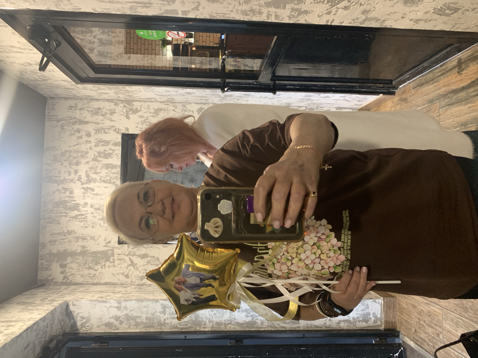 A woman with short blonde hair and glasses, wearing a brown t-shirt and dark pants, stands in a hallway and takes a selfie with her phone. She is holding a star-shaped gold balloon adorned with a photograph of two older people and a bouquet of pink and white flowers attached to it by a white and gold ribbon. The hallway has textured gray and white walls and a wooden floor. To the right of the woman, another woman with reddish-pink hair, wearing a white jacket, is visible in the reflection of a glass door. The scene is likely indoors, possibly in a public or commercial building, given the presence of the glass door with a visible sign inside. The overall impression is a casual moment captured through a selfie.