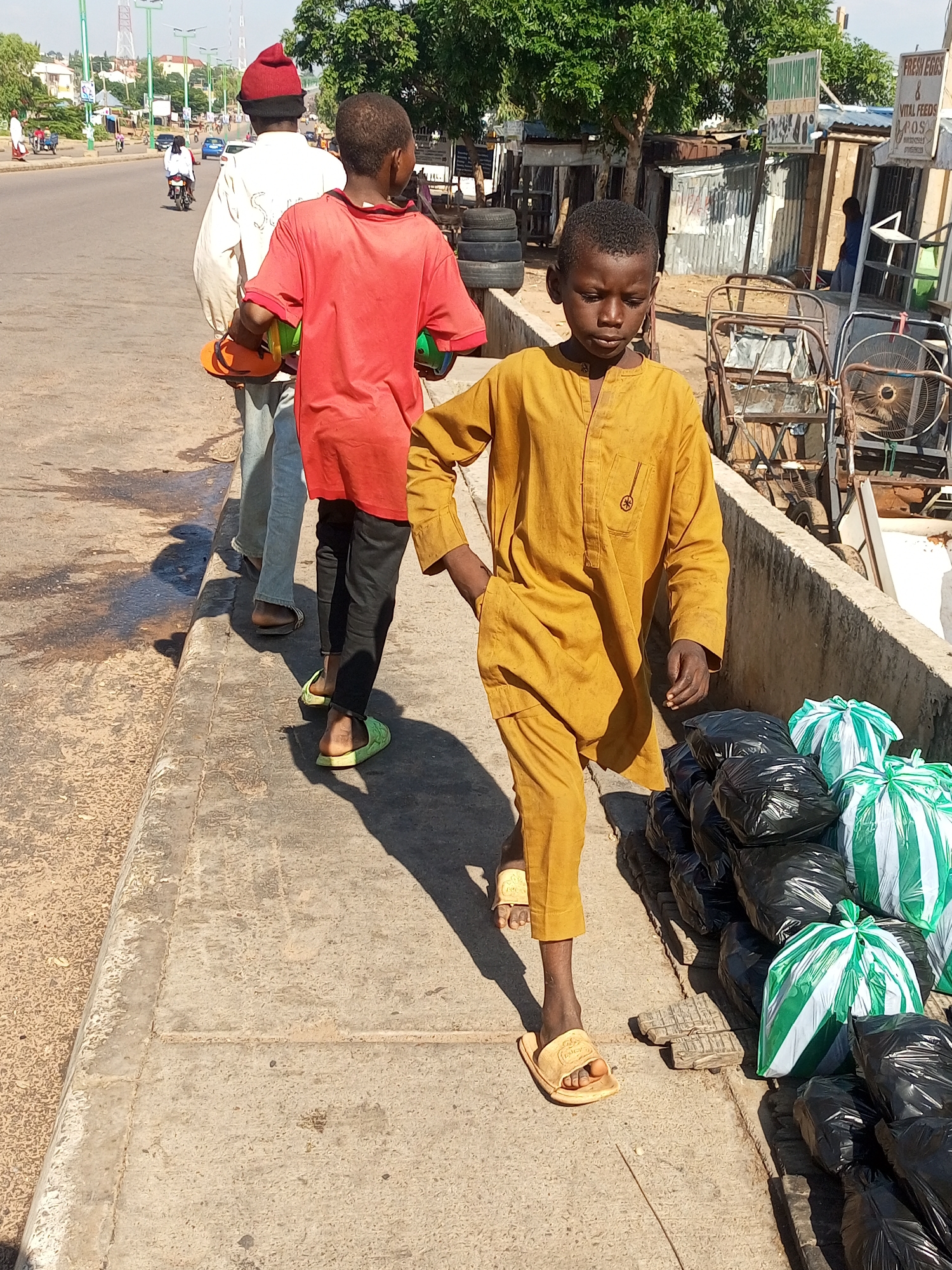 A medium shot captures a sunny outdoor scene on a concrete sidewalk with a paved road to the left and a row of stalls and businesses to the right. In the foreground, a young boy wearing a yellow tunic and matching pants walks towards the right, his head down. He is wearing light-colored sandals. To his left, another boy in a red t-shirt and black pants walks away from the camera, his head also down. He wears green sandals and carries two colorful objects in his left arm. Behind him, a man in a cream-colored robe and red beanie walks in the same direction. He carries orange and green objects in his right arm. Several black trash bags and some green and white striped bags are piled to the right of the foreground boy. In the background, traffic is visible on the road, including cars and motorcycles. Trees and buildings line the other side of the road. A few signs are visible on the stalls, including one that reads "FRESH EGGS & VITAL FEEDS." The overall impression is a bustling street scene in a Nigerian city.