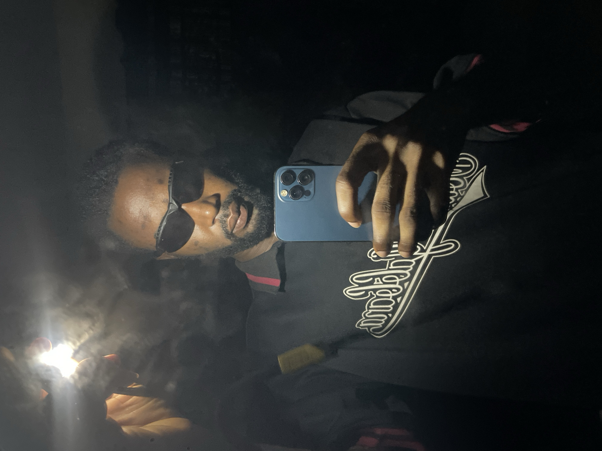 The image displays an adult male, with dark skin, a beard, and dark curly hair, engaged in taking a selfie within a reflective surface in a dimly lit environment, specified as Lokoja, Nigeria. He wears dark sunglasses and a dark polo shirt featuring a mirrored white graphic on the chest. His right hand holds a blue smartphone, identifiable by its visible triple camera array. His left hand is positioned to the upper left, holding and operating an external, highly luminous light source, which generates significant lens flare and intense highlights. The scene's illumination primarily stems from this external source and the smartphone's flash, casting the majority of the background into deep shadow, with minimal discernible environmental features.