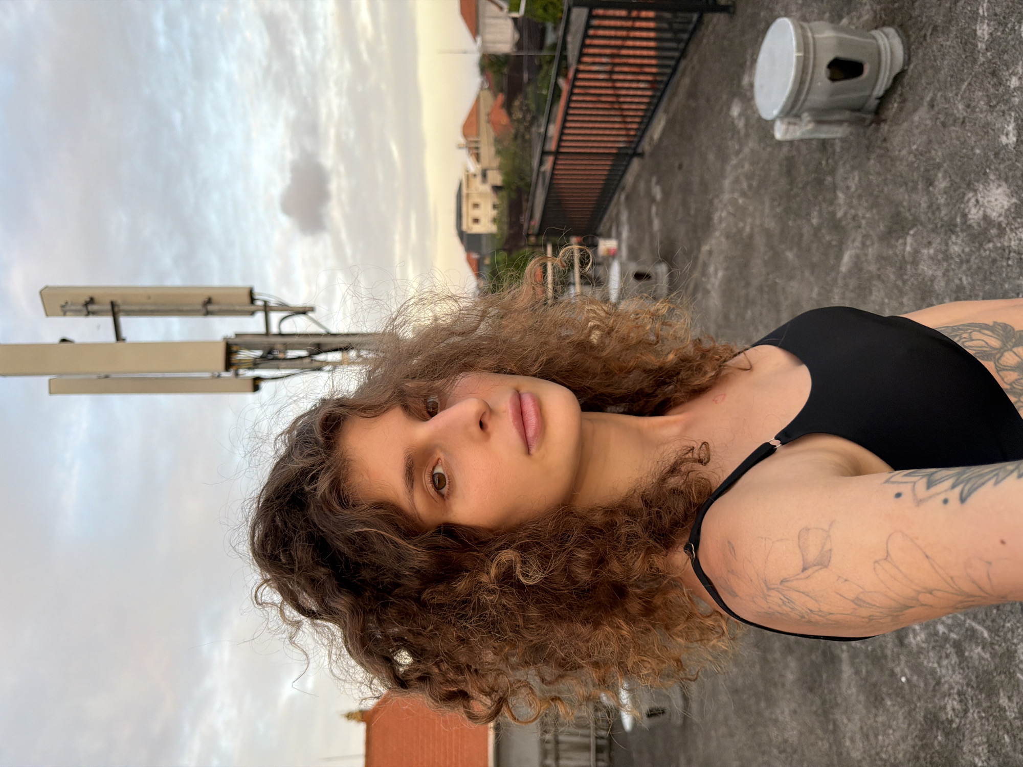 A medium shot, angled slightly, captures a woman with brown curly hair and tan skin. She is wearing a black top with thin straps. A light-colored tattoo is visible on her left arm.
In the background, there are buildings with orange roofs, typical of the region. There's also a metal railing and a gray, cylindrical object with holes. A communications tower stands to the left.
The setting is North Kuta, Indonesia, on a rooftop or elevated surface. The sky overhead is a mix of cloudy gray and pale yellow.