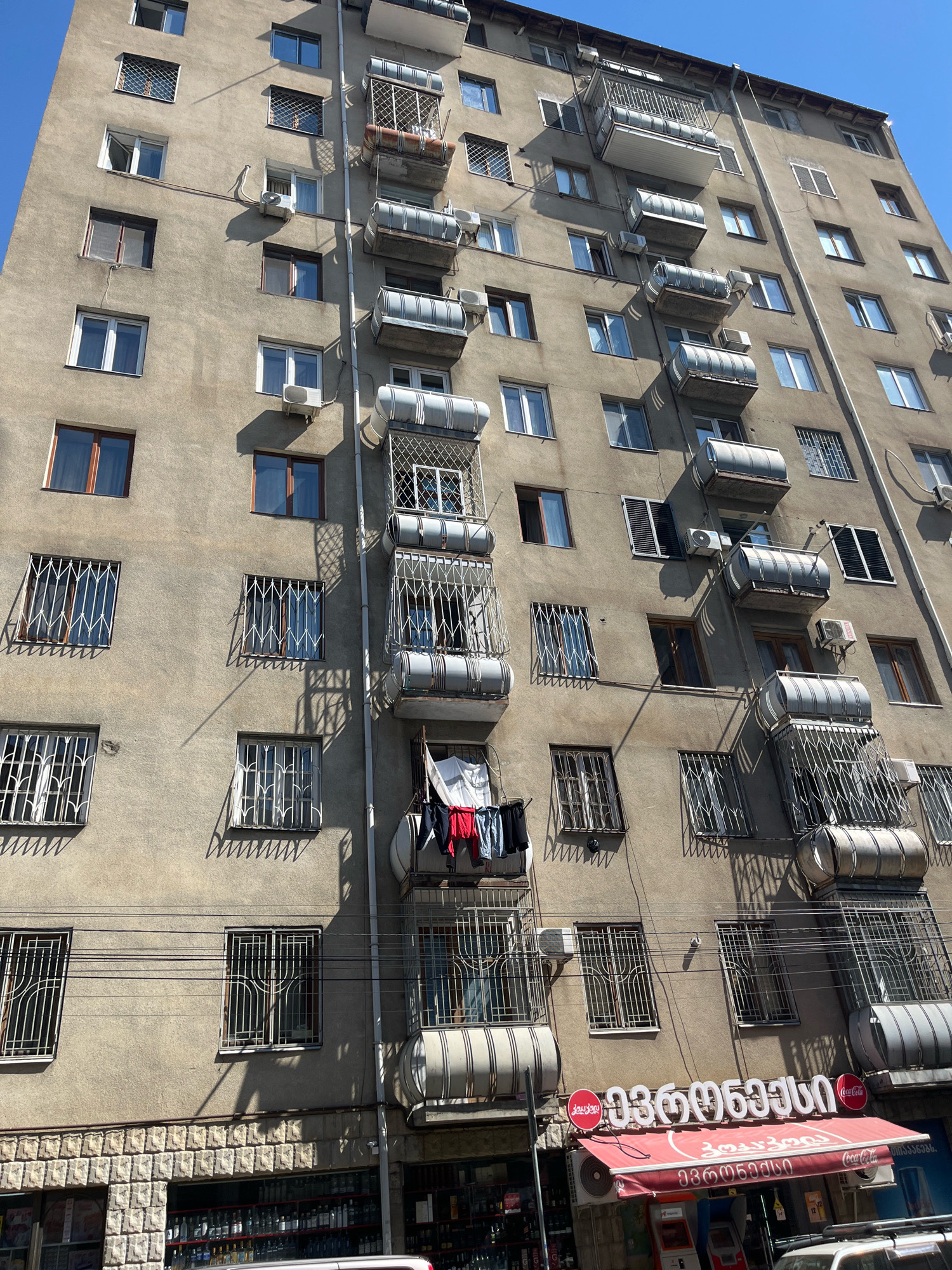 The image shows a multi-story apartment building with a weathered facade, likely in Tbilisi, Georgia, given the Georgian text on the awning below. The building has a symmetrical arrangement of windows and balconies, many of which are enclosed or have decorative metalwork. Several air conditioning units are visible on the exterior walls. Clothes are hanging to dry on one of the balconies. The lower portion of the building features a storefront with a red awning and signs in Georgian, indicating a retail establishment, possibly a grocery store or small supermarket. Electrical wires are strung across the facade. The sky is clear and blue, suggesting a sunny day.