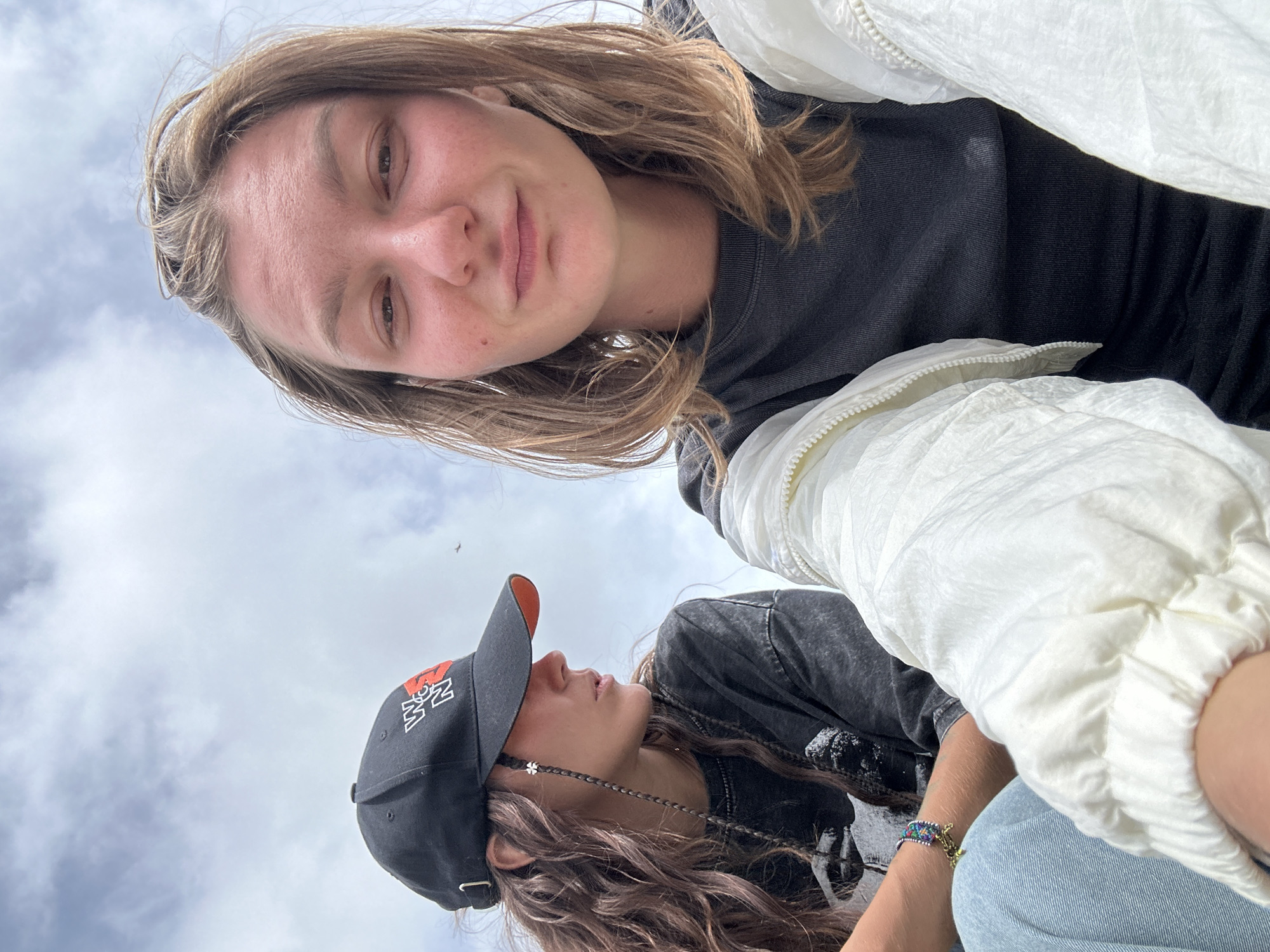 Two individuals are outdoors, seated against a sky with scattered clouds. The person in the foreground is looking directly at the camera, with their face close-up and shoulders visible. They are wearing a dark grey t-shirt and a white puffer jacket. The person in the background, to the left, is wearing a black baseball cap with an orange brim and a graphic t-shirt, and is looking off to the side. They have long, wavy brown hair with a thin braid hanging down, adorned with a small white charm. The background is dominated by the sky. No specific landmarks or details indicating the location in Bắc Yên, Vietnam, are visible.