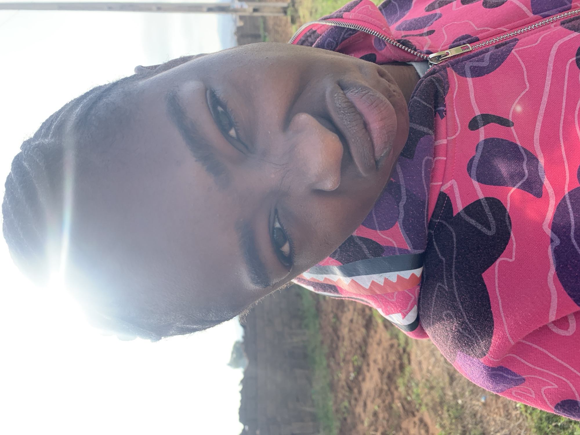 A rotated portrait-style image captures a person with dark skin looking directly at the viewer. The individual is wearing a distinctive hooded sweatshirt that is predominantly pink, with large purple and black abstract or camouflage-like patterns. A metallic zipper runs down the front of the hoodie. On the cuff or lower sleeve, a graphic design resembling a shark's mouth with white, orange, and black elements is visible.
The subject's head is tilted, and their dark hair, possibly braided or very short, is visible. Their eyes are dark, and their lips appear full. A bright lens flare from strong sunlight obscures the upper left portion of the frame, falling across the subject's forehead and hair, suggesting the sun is low in the sky—either morning or late afternoon.
The setting is outdoors, appearing to be a rural or undeveloped landscape in Unguwar Saya, Nigeria. The background shows areas of brown earth or dirt and sparse green vegetation. Faint structures, possibly utility poles or distant buildings, are visible further back under a bright, hazy sky. No visible text is present in the image.