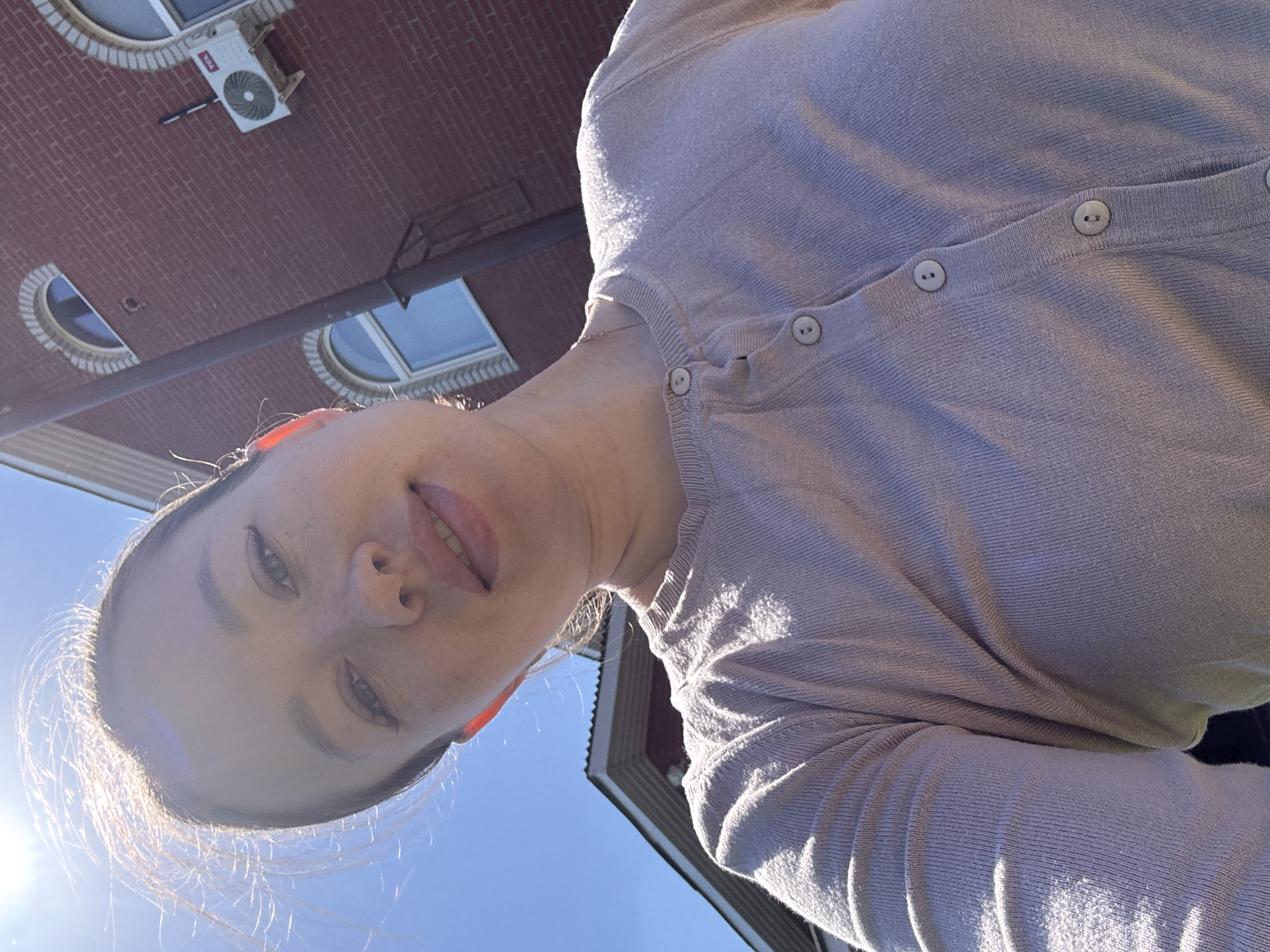 A woman's face is captured from a low angle, tilted, with the sky visible in the background. She has fair skin and is wearing a light-colored cardigan. Behind her, the roof of a building is visible, constructed with red bricks and featuring arched windows. A white air conditioning unit is mounted on the building. The photo was taken in Ферма, Russia.