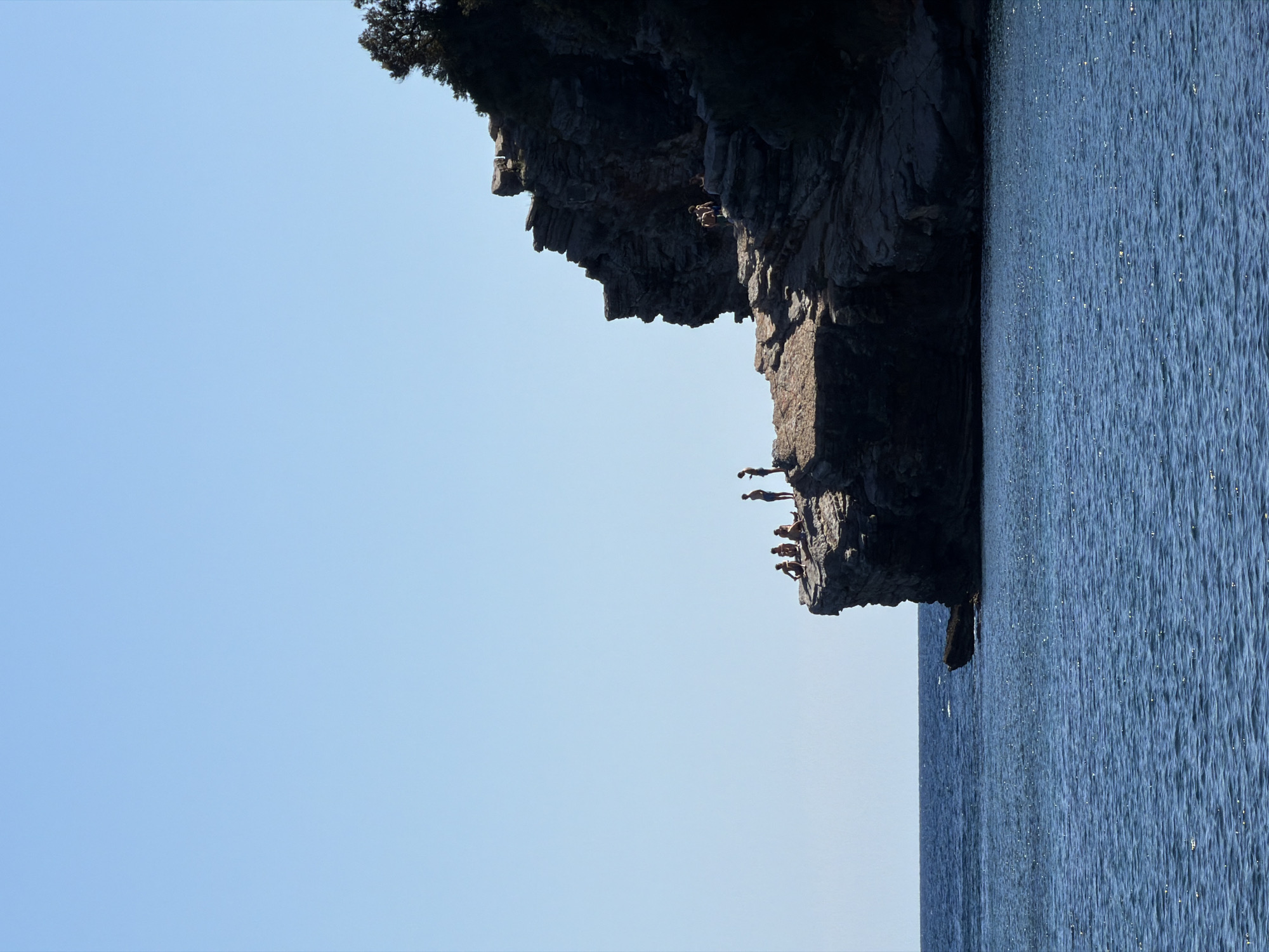This image captures a dynamic coastal scene in Budva, Montenegro, primarily featuring a tall, rugged rock cliff meeting the sparkling sea under a clear blue sky. The photograph, presented rotated 90 degrees clockwise, shows a vast expanse of blue sky on the left and the deep blue, rippled water on the right, with the dark, textured cliff face cutting diagonally through the center.
When viewed in its natural orientation, the scene depicts a group of people engaged in cliff jumping. Several small figures are visible on various ledges of the imposing rock formation. Approximately 6-8 individuals are gathered on the lower parts of the cliff, appearing to be observing or awaiting their turn. Higher up on the cliff, one person is distinctly captured in mid-air, in the act of jumping from the rock face towards the water below.
The setting suggests a bright, sunny day, likely midday or early afternoon, given the intensity of the blue sky and the numerous points of sunlight glinting off the water's surface, indicating a clear, cloudless sky. Some green vegetation, possibly trees or bushes, can be seen at the very top edge of the cliff, adding to the natural landscape. The image conveys a sense of adventure, excitement, and anticipation associated with the activity of cliff diving. There is no visible text in the media file.