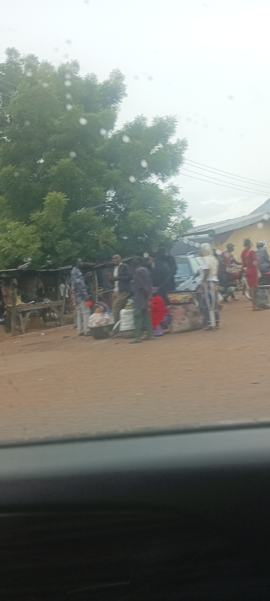 The image, taken from inside a vehicle, shows a street scene in Gwadanye, Nigeria. A group of people are gathered near a small van, possibly a commercial vehicle. Some people are standing near the van, while others are on motorbikes. There are rudimentary stalls or shops nearby. The road is unpaved. The environment is overcast, with trees in the background.