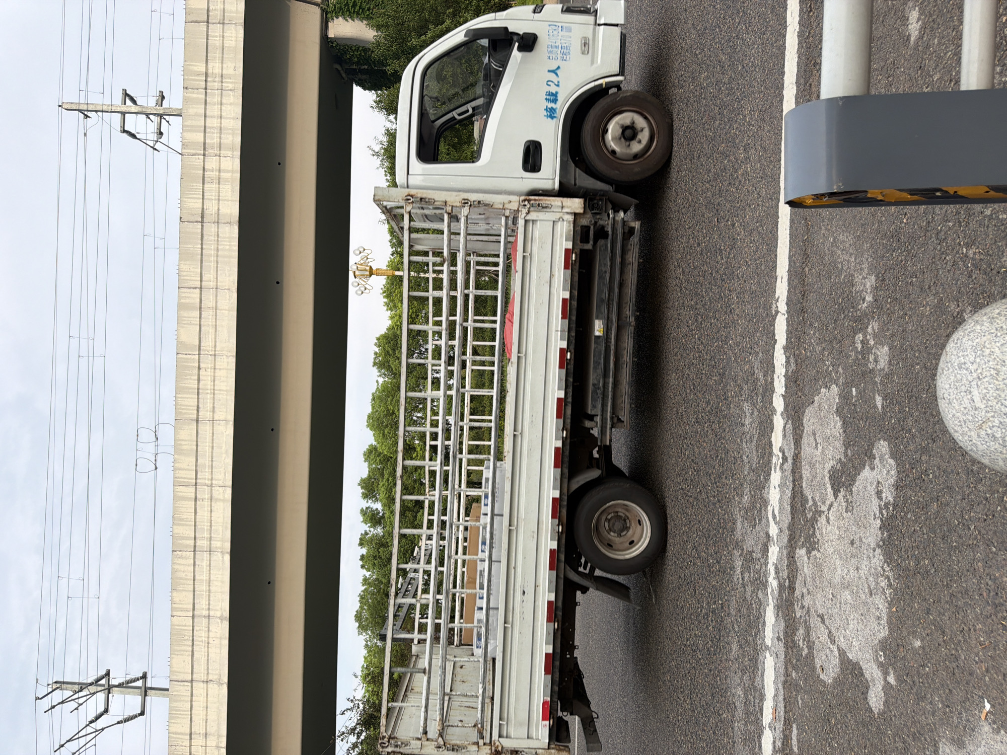 A white, flatbed truck with a metal cage bed is driving on a paved road in Jiangjin, China. The truck is carrying several boxes in its bed. The truck has Chinese characters on its side door. An elevated concrete structure, possibly a highway overpass, is visible above the truck. Trees and foliage are present behind the truck. The sky is overcast. The road has a white painted line and some reflective markers. The scene is captured from a side profile perspective.