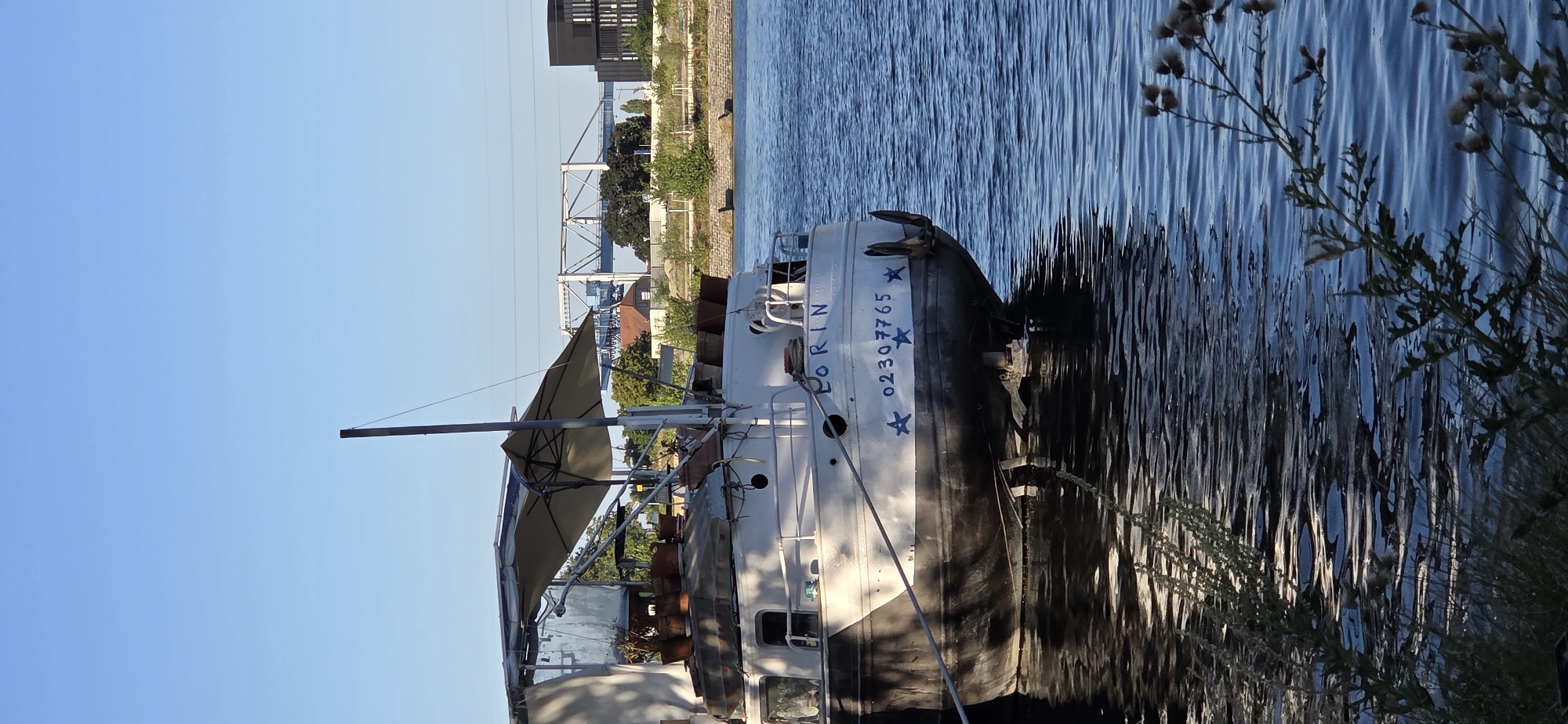 This is a photograph taken from a slightly low angle, capturing a white boat named "LORIN" on a body of water. The boat is moored near a bank with some greenery. The boat has several potted plants on its deck, and a large umbrella is deployed for shade. On the stern of the boat, the name "LORIN" is visible in blue lettering, along with a phone number "02307765" and several blue stars. The water reflects the sky and the boat. In the background, there is a bridge, some buildings, and more greenery, suggesting an urban or semi-urban environment. The sky is clear and blue. The overall lighting suggests it is daytime, likely late afternoon due to the angle of the sunlight. The scene appears to be peaceful and stationary. While the image is not explicitly identified as being in Strasbourg, France, the presence of a bridge and urban elements is consistent with many waterfront areas in such cities.