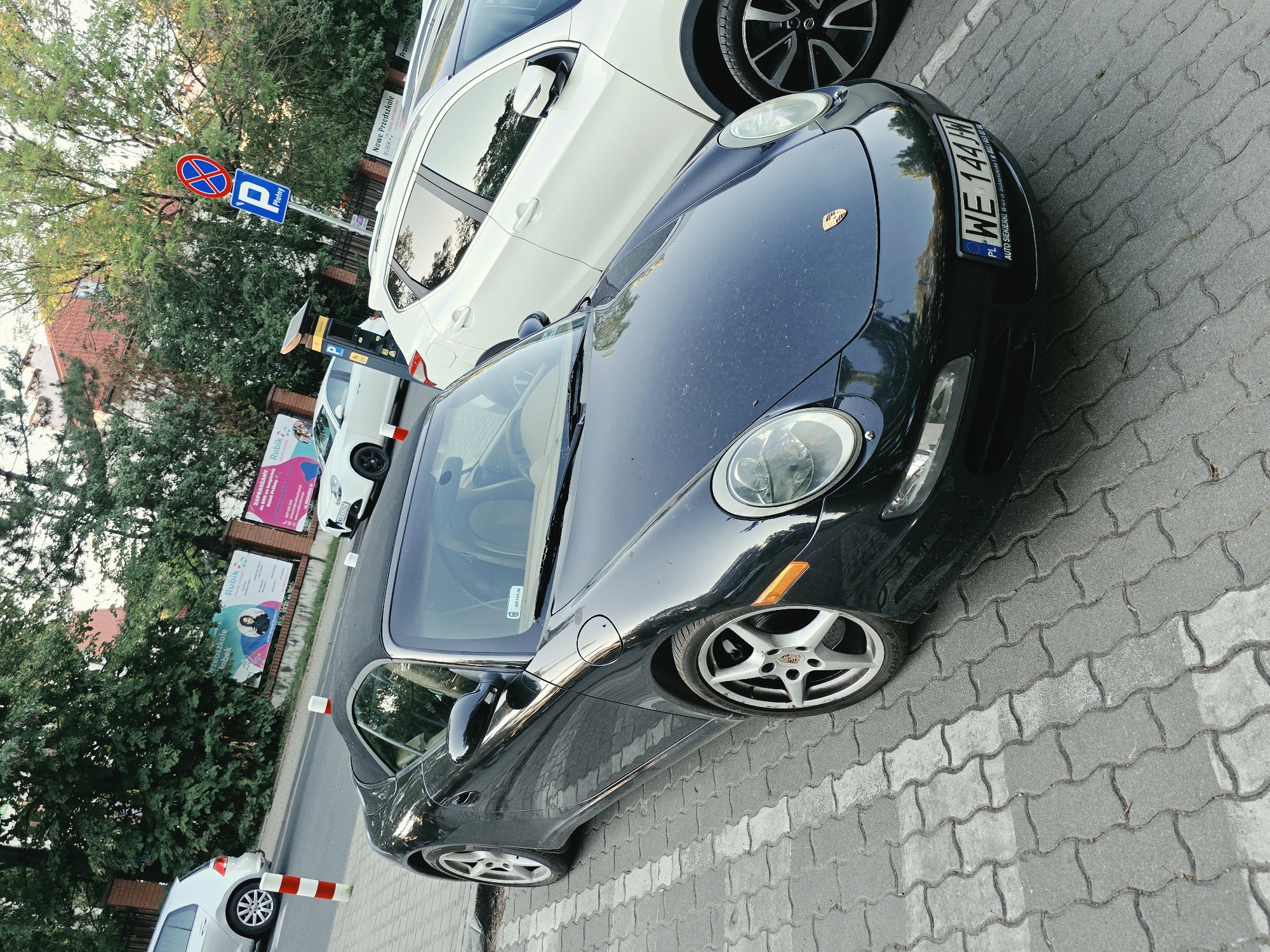 The image shows a street scene in Warsaw, Poland, with several cars parked on a paved lot. A dark Porsche sports car is prominently featured in the foreground, its front end angled towards the viewer. Next to it, a white SUV is parked. In the background, trees and buildings are visible. A parking sign with a "P" symbol and a red circle with a diagonal line indicate paid parking. There are also advertisements on a fence, including one for "Rubik Mobek Przedszkole" (Rubik Mobek Kindergarten) and another for "Nowe Przedszkole RUBIK" (New Kindergarten RUBIK). The license plate of the Porsche reads "WE 144JM". The lighting suggests it is daytime, likely late afternoon or early evening, given the soft light. The scene appears to be a residential or commercial area, with the parked cars and advertisements suggesting a public space. There are no people visible in the image.