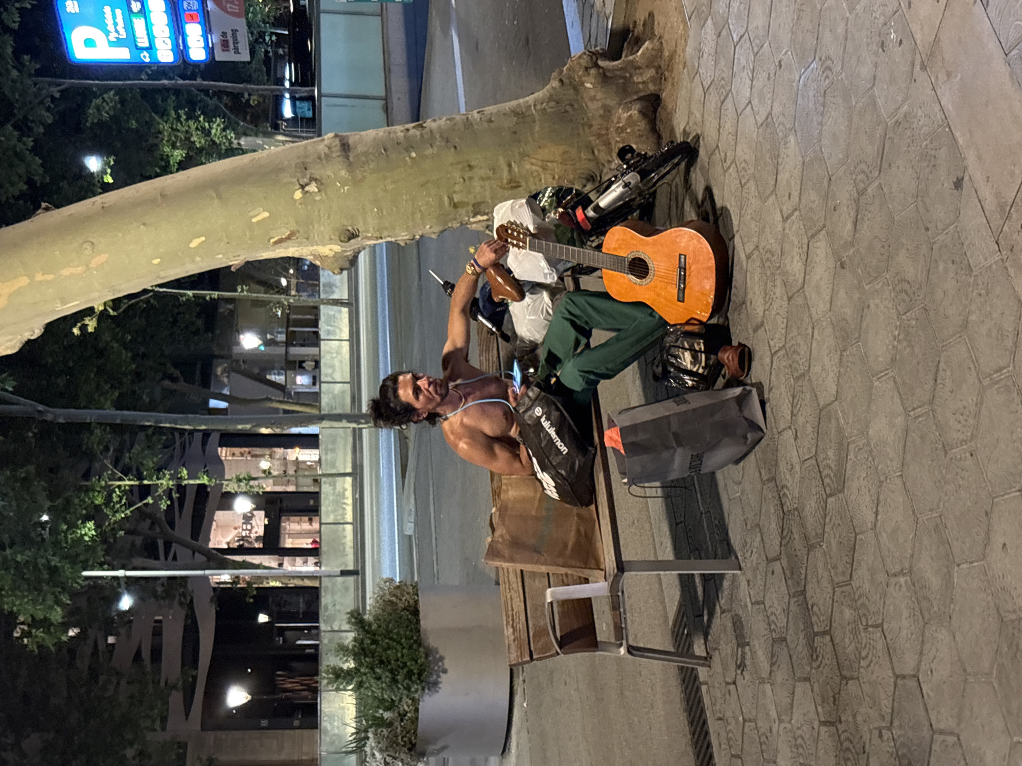 A shirtless man with long dark hair tied in a bun sits on a bench on a Barcelona street. He wears green pants and has an orange guitar resting on his lap. The man has a backpack and a bag next to him. Behind the bench is a large tree trunk, and in the background, there are buildings, trees, and streetlights. A bicycle leans against the tree trunk. A parking sign is visible in the upper left corner. The ground is paved with hexagonal stones.
