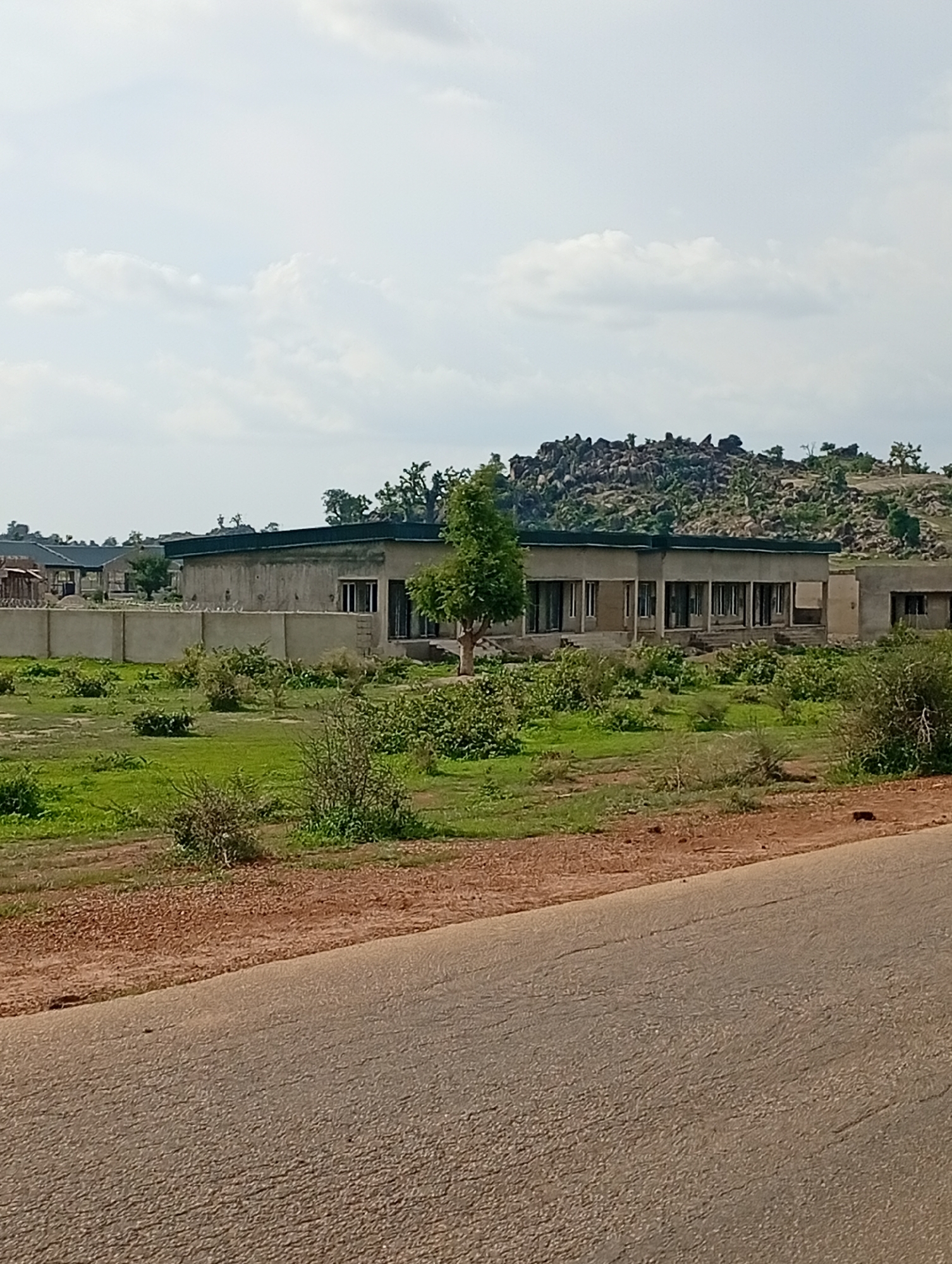 A low-angle, medium shot captures a view of an unfinished building complex with a wall surrounding it. The building, constructed from concrete and adorned with a dark roof, features numerous openings that appear to be windows or doorways. A single tree stands tall next to the structure, its branches reaching towards the sky. The surrounding environment is characterized by lush green grass and scattered bushes, suggesting a rural or semi-rural setting, possibly in the city of Shuwa, Nigeria. In the distance, a rocky outcrop with sparse vegetation rises against a cloudy sky. A paved road occupies the foreground of the image, its surface textured and showing signs of wear.