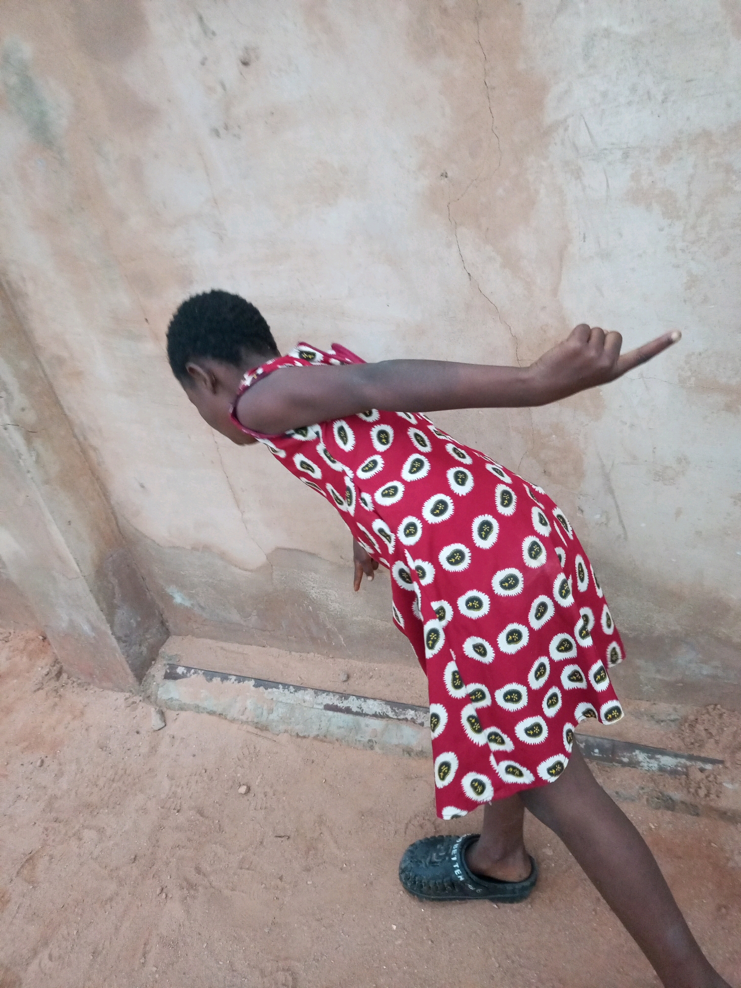 In Tempure, Nigeria, a young person wearing a red dress with a white and black pattern stands on sandy ground near a textured, light-colored wall. The person is leaning forward with their right arm extended backward and slightly upward, hand open with index finger extended. Their left arm is bent downwards. They are wearing black clogs on their feet. The environment appears to be outdoors, possibly a courtyard or alley, with the wall exhibiting some cracking and staining.