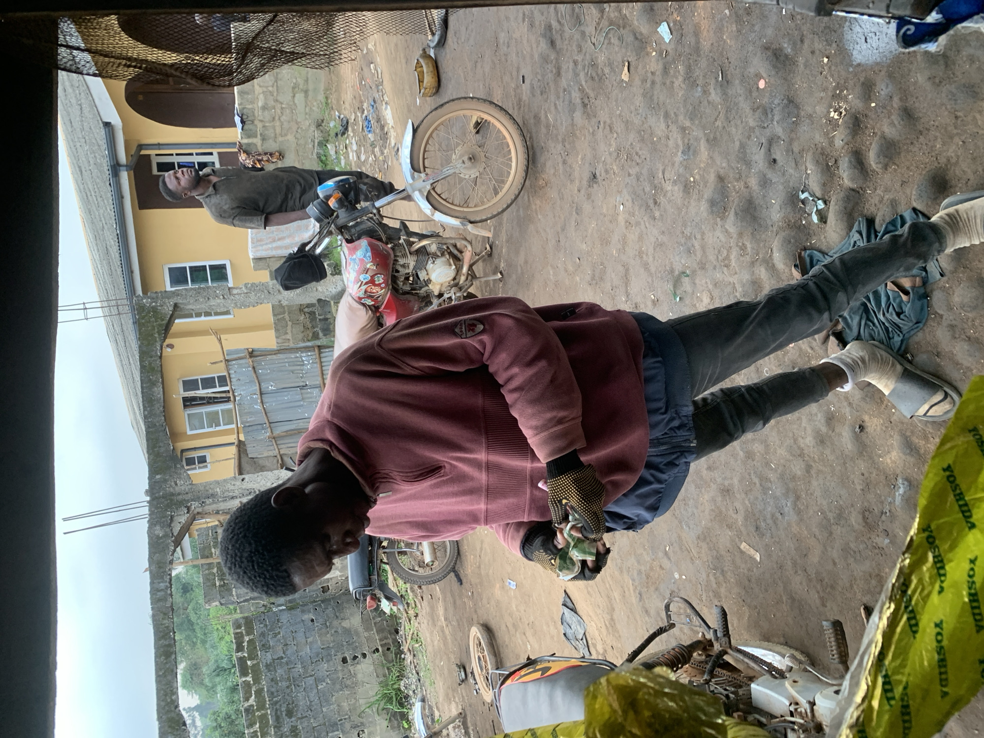 In Shagamu, Nigeria, a young man wearing a maroon sweatshirt and dark jeans stands in an outdoor workshop, examining several small green objects in his gloved hands. He is focused on the items, which appear to be rolled-up bills or small packages. Next to him, a red motorcycle is parked, its front wheel turned to the side. In the background, another man in a dark t-shirt stands near a yellow building. The ground is dirt and gravel, with scattered debris. Other motorcycles and parts are visible in the immediate vicinity, suggesting a mechanic's or repair shop environment.