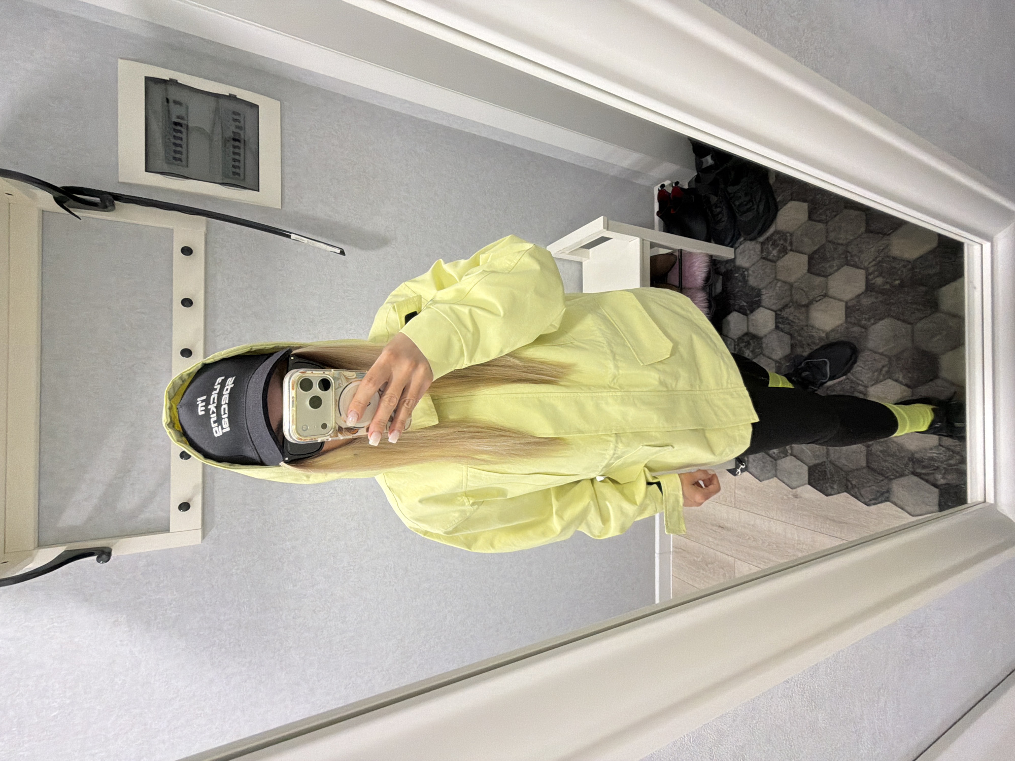 A person is captured in a mirror selfie, standing in what appears to be an entryway or dressing area within a home in Votkinsk, Russia. The image (originally rotated 90 degrees counter-clockwise) shows the individual from the chest down, holding a smartphone that obscures their face. They are wearing a bright, light yellow hooded jacket, black leggings, and black sneakers with neon yellow-green accents, possibly visible socks. A black baseball cap is worn on their head, displaying the reversed text "BALENCIAGA." The person has long blonde hair and neatly manicured fingernails with French tips.
The reflected background reveals a modern interior. The walls are a textured light grey. On the left, a white-framed electrical panel or fuse box is mounted on the wall, below which is a white coat rack with black knobs. The floor features a distinct pattern of hexagonal tiles in varying shades of grey and black, transitioning to lighter wooden planks on the right side of the image. A white shelving unit or shoe rack on the right holds several pairs of dark footwear and a pink, fluffy item. The scene is brightly lit, indicative of indoor lighting. The primary activity is the subject taking a self-portrait in the mirror, likely to showcase their outfit.