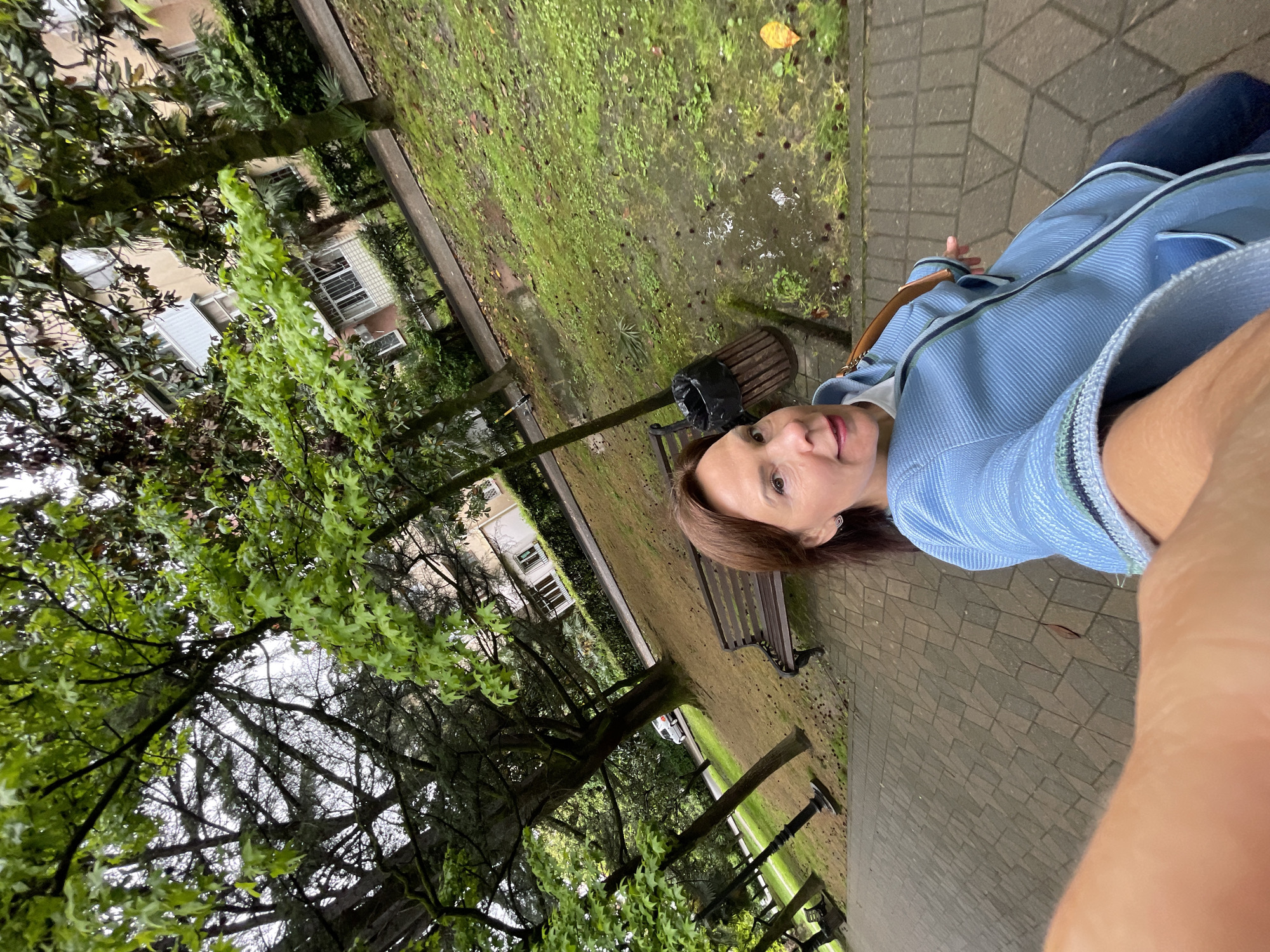 A woman with brown hair is taking a selfie on a paved path in a park-like setting. She is wearing a light blue jacket and jeans. Behind her, there's a patch of green grass with puddles and what looks like moss. Benches and a trash can are visible along the path. Trees with green leaves and some buildings in the background can be seen. It is possibly Sochi, Russia, but more context is needed for confirmation.