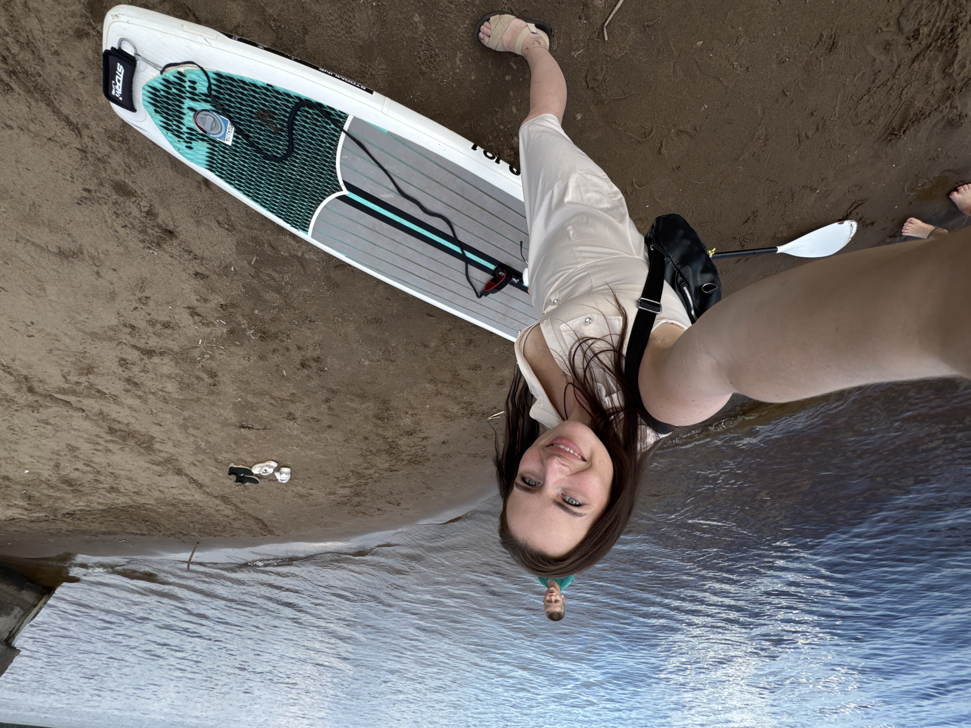 This is an inverted, wide-angle selfie taken by a young woman on a sandy shore next to a body of water. The woman is the primary subject, smiling at the camera. She is wearing a light-colored dress and sandals. Beside her lies a white stand-up paddleboard with grey and teal accents, a black bag, and a paddle. In the background, there are some shoes and a small child in the water. The scene appears to be a beach or lakefront. The lighting suggests it could be daytime. The word "STORM" is visible on a black tag attached to the paddleboard. The overall mood seems happy and relaxed, indicative of a leisurely activity on a summer day. The location is Chaikovsky, Russia.
