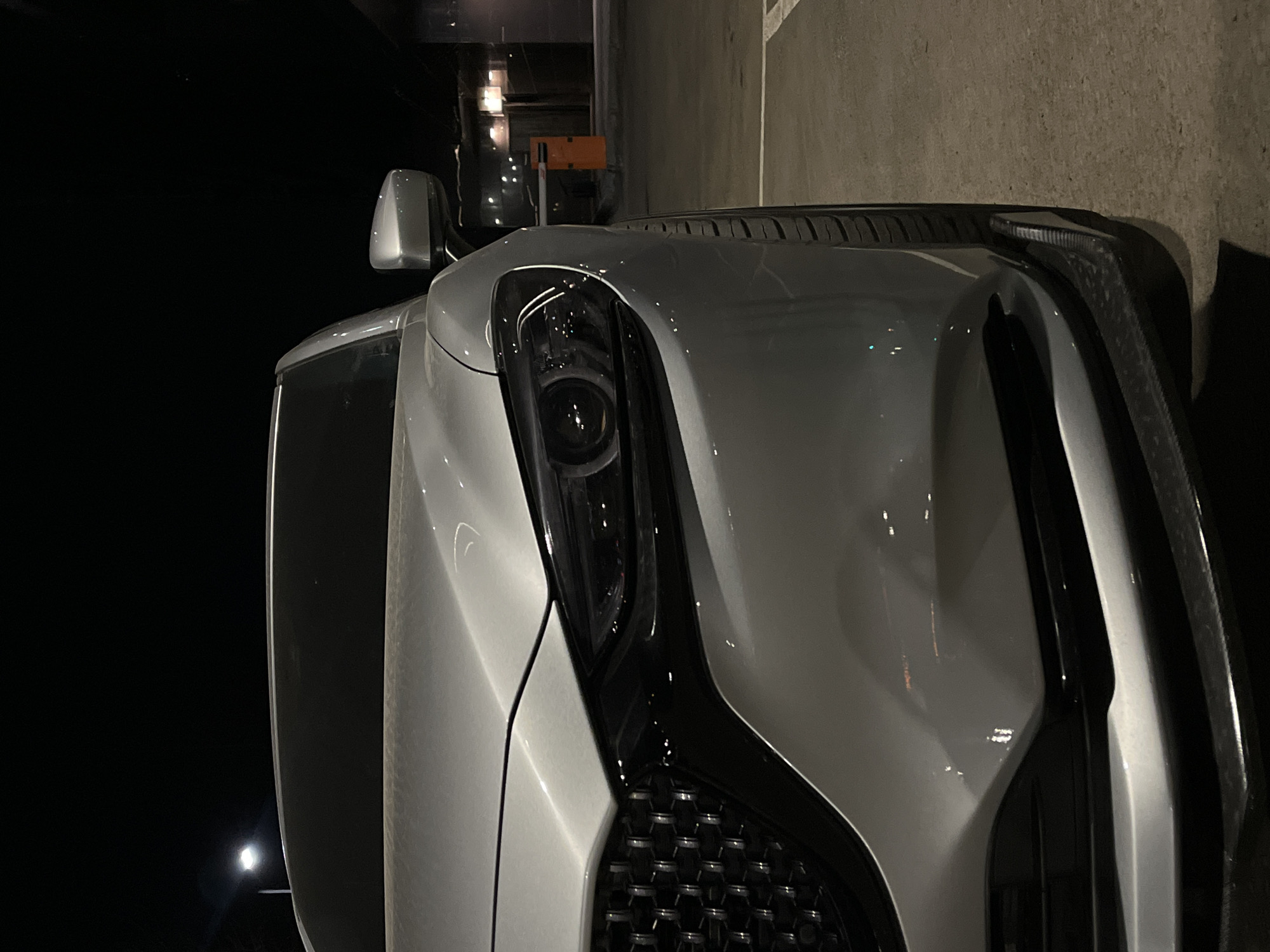 The image shows the front of a silver car at night. The headlight is prominent, and a side mirror is visible. The car is parked in what appears to be a parking lot or near a building. The background is dark, but there are some lights in the distance. There is no indication of the location being in Krasnoyarsk, Russia, or any other specific place, nor is there any evidence of people, actions, or interactions in the scene. The image focuses primarily on the vehicle and its immediate surroundings.
